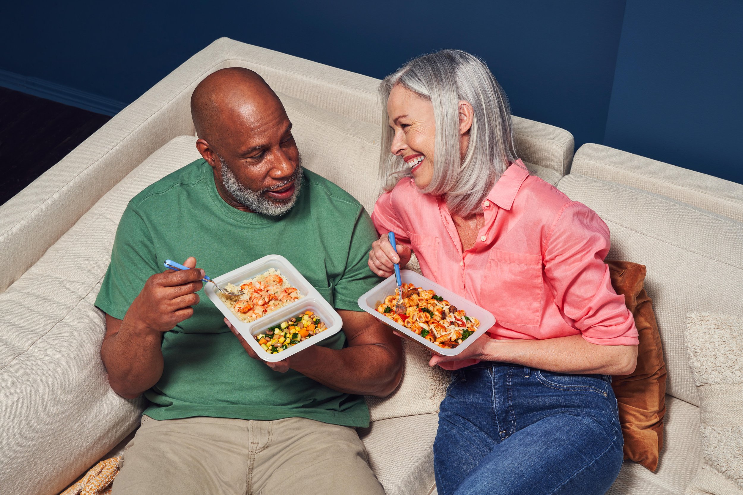 A mid-50s couple laughing and enjoying fully-prepared Freshly meals on the couch.