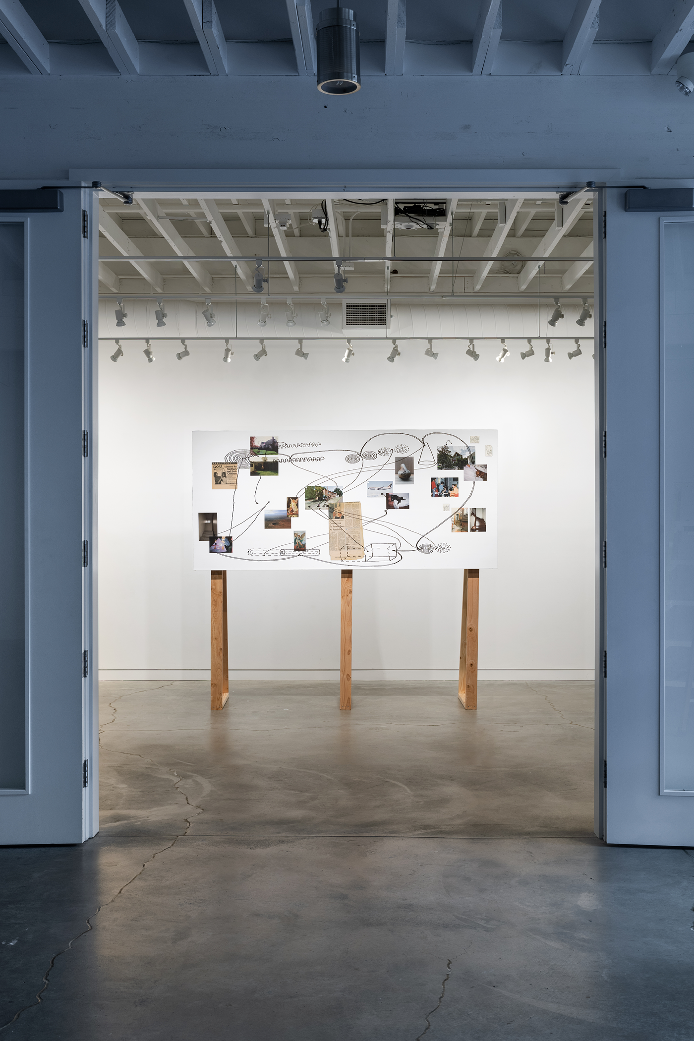 A vertical white board on three wooden legs displays scattered snapshots, newspaper clippings, and postcards floating amongst one another and encircled and overlapped by line drawings of geometric shapes, swirls, and snaking lines.