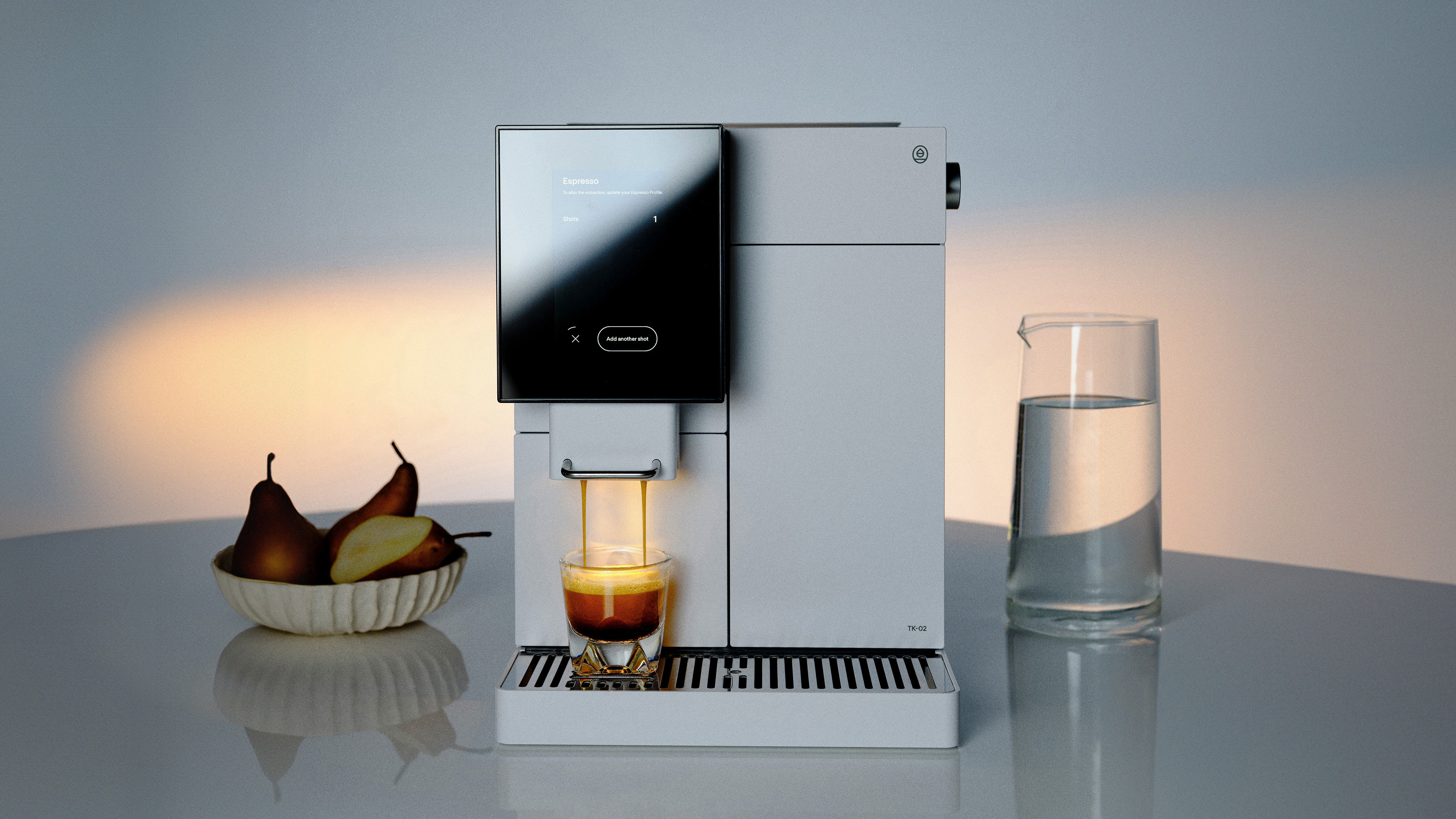 A white TK-02 on a blue table pouring an espresso shot next to a bowl of pears and a glass of water