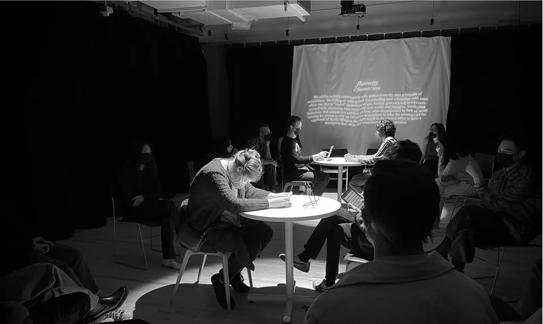 Two conversations happen in the center of the room as an audience watches, one participant has a limited vocabulary of certain words in front of them, one asks questions
