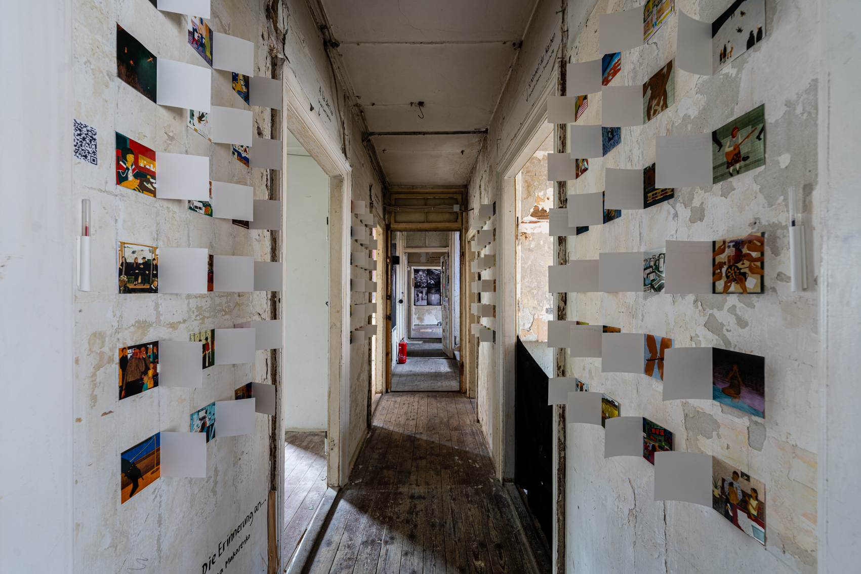 Bild 1 zu Die Erinnerung an..: Das Bild zeigt den Blick in den Gang der WG Anima. An den teils abbl&auml;tternden wei&szlig;en W&auml;nden h&auml;ngen &uuml;ber 100 aufklappbare Illustrationen in Reihen &uuml;bereinander. Diese sind etwa 10 auf 10 Zentimeter gro&szlig; und zeigen Ausschnitte aus dem Leben der K&uuml;nstlerin. Auf die aufklappbare wei&szlig;e Au&szlig;enseite k&ouml;nnen Besucher*innen schreiben, welche Erinnerung die Illustration in ihnen weckt. 