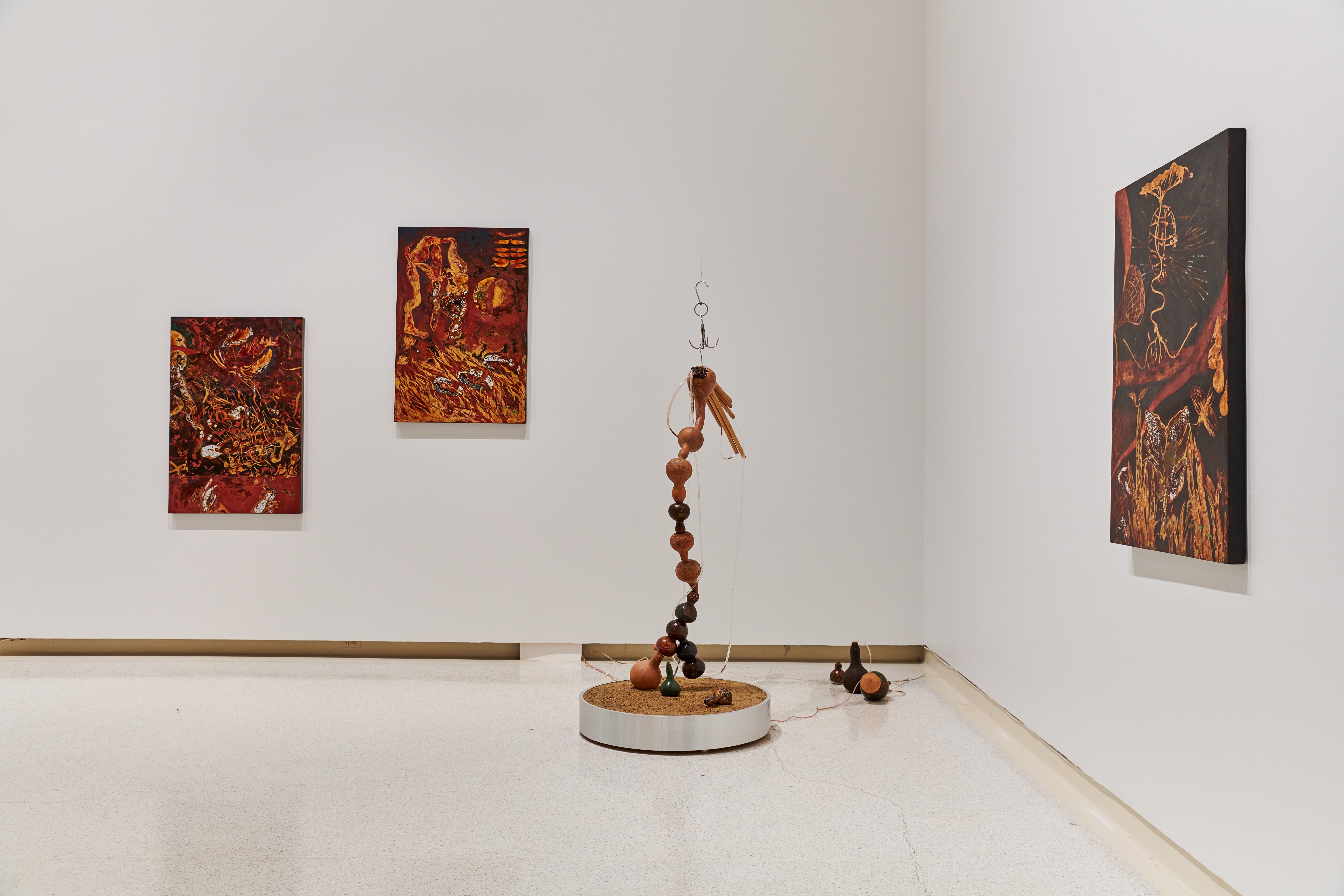 In the corner of a white wall gallery space there are three hanging paintings that resemble each other, two directly in front, and one to the right, and in between is a floor standing abstract sculpture that resembles a larger than life fishing lure.