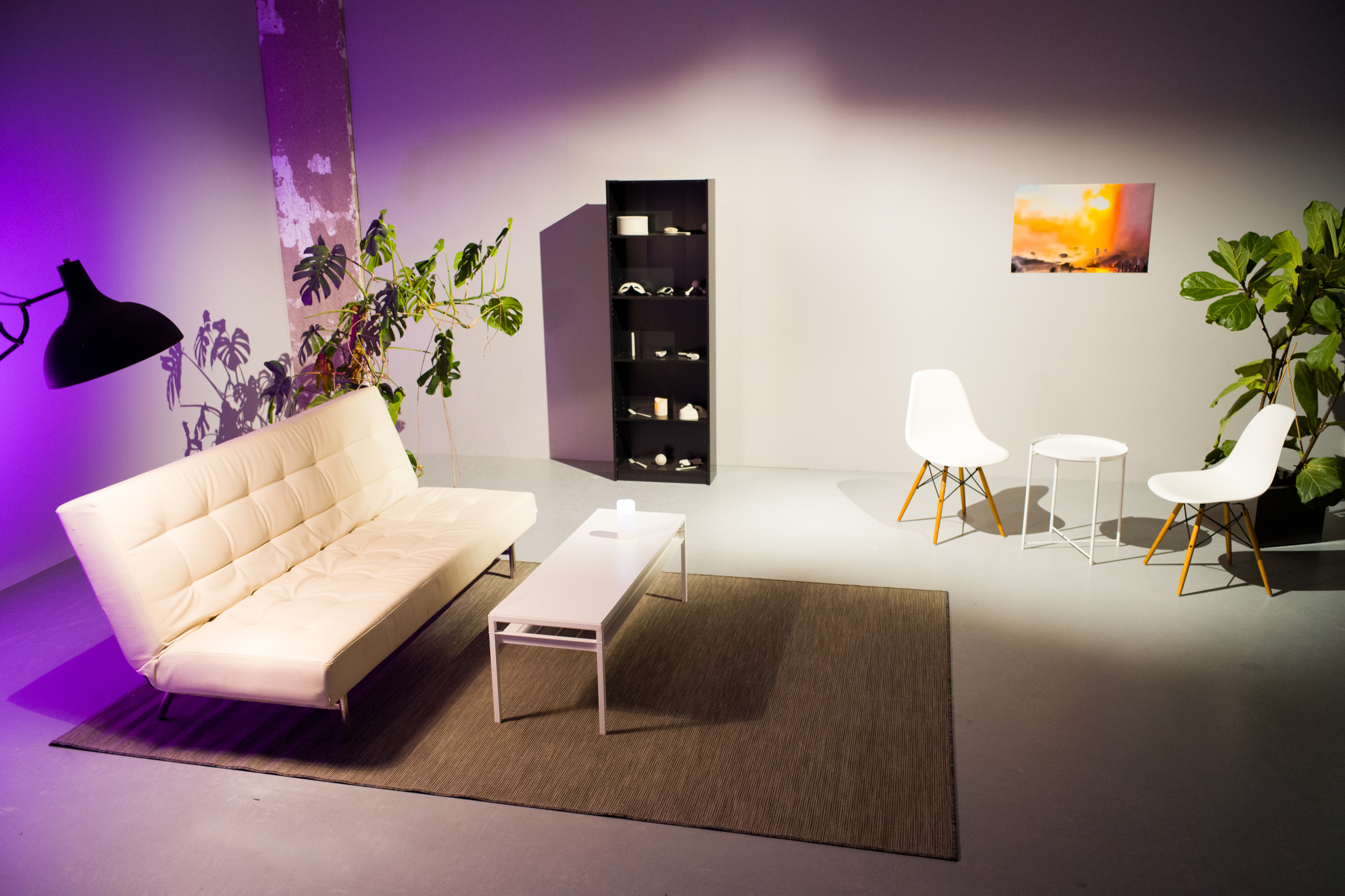 image of a room with a white couch, white table, two chairs, another white table, a lamp, two plants, a book shelf, a landscape photograph and a rug.