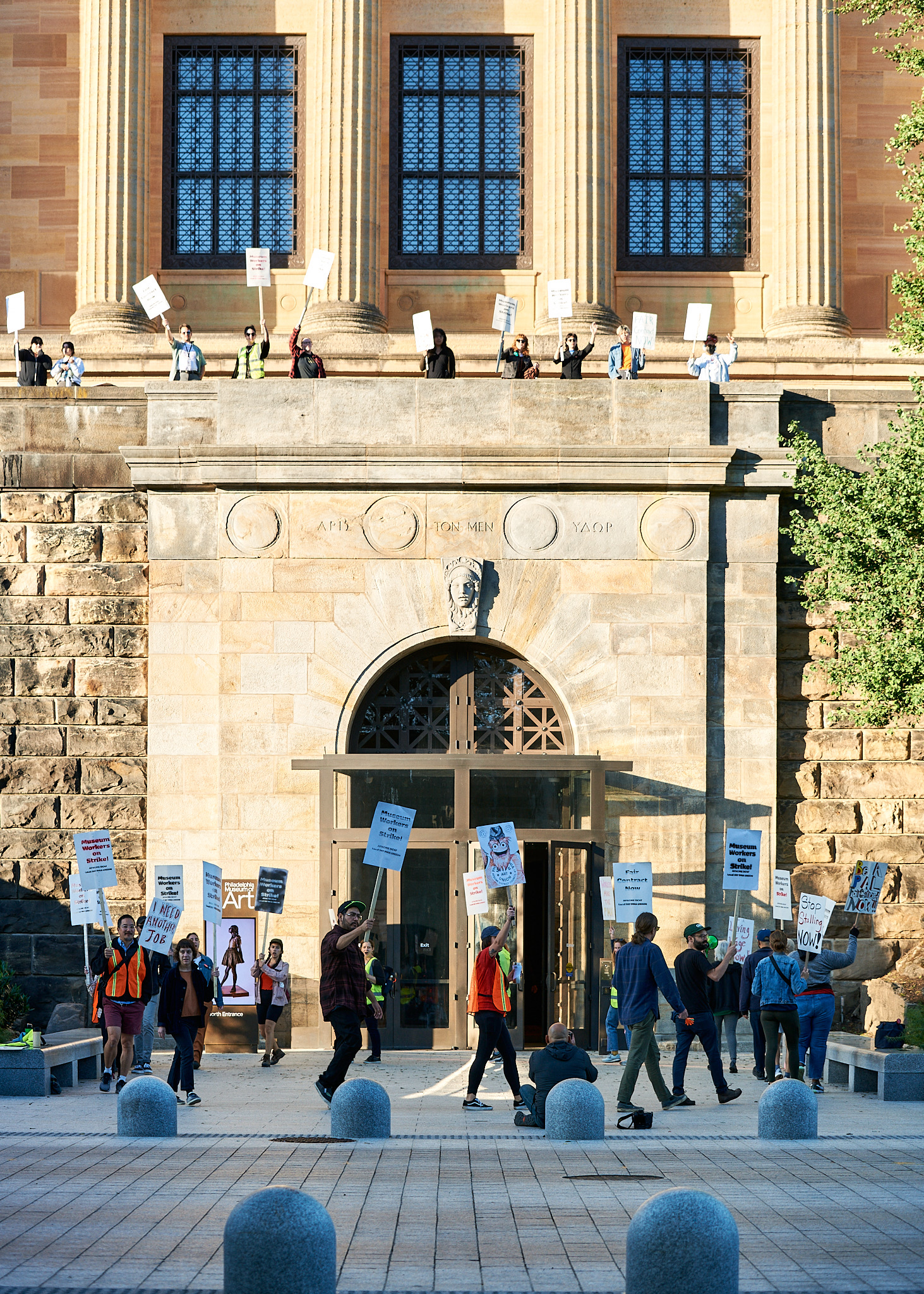 Color photo of a group of many striking workers holding picket signs in front of an entrance to the Philadelphia Museum of Art, and moving in a circle, as if seen from the street. Above can be seen even more strikers with signs on a balcony overlook.