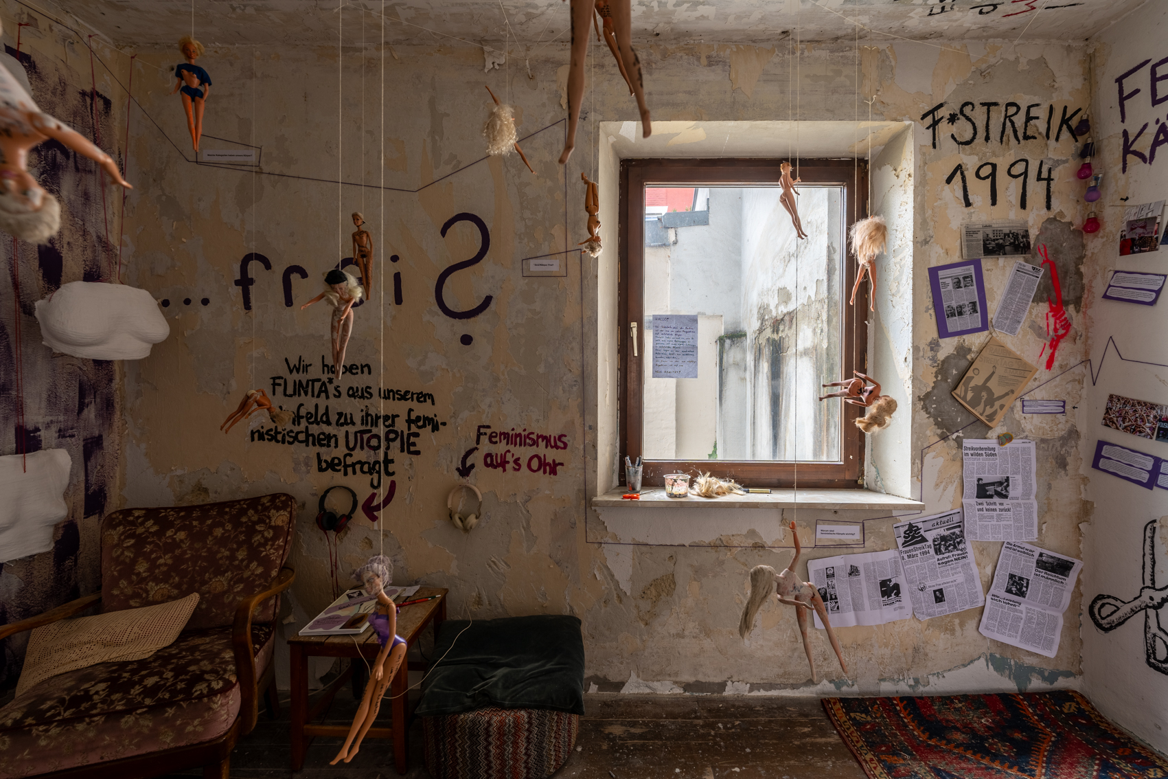 Bild 3 zu Patriarchat abnippeln: Blick auf die Wand gegen&uuml;ber des Eingangs. Mit einem Fenster in der rechten Wandh&auml;lfte. Von der Decke h&auml;ngen Barbiepuppen. Der Putz an den W&auml;nden bl&auml;ttert teilweise ab. Links steht der Sessel. Rechts vom Fenster h&auml;ngen einige fr&uuml;here Zeitungsausschnitte zu Feministischen Streiks. Dar&uuml;ber steht F*Streik 1994.