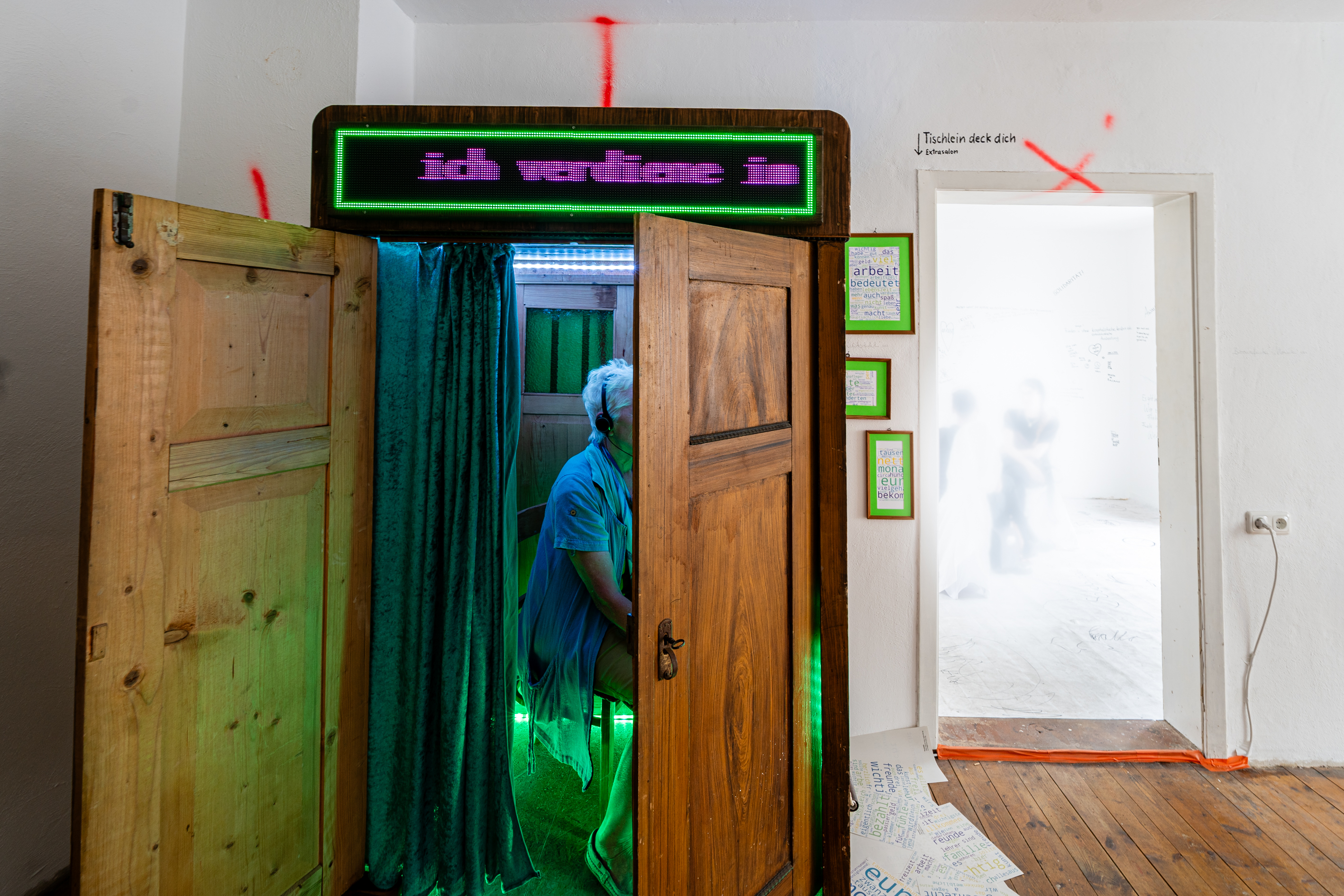 Bild 2 zu Un&ne;gleich: Ein Holzschrank mit ge&ouml;ffneter Doppelt&uuml;r. Oberhalb der T&uuml;r befindet sich eine digitale Anzeigetafel, die anzeigt, was die Person im Inneren des Schrankes antwortet.