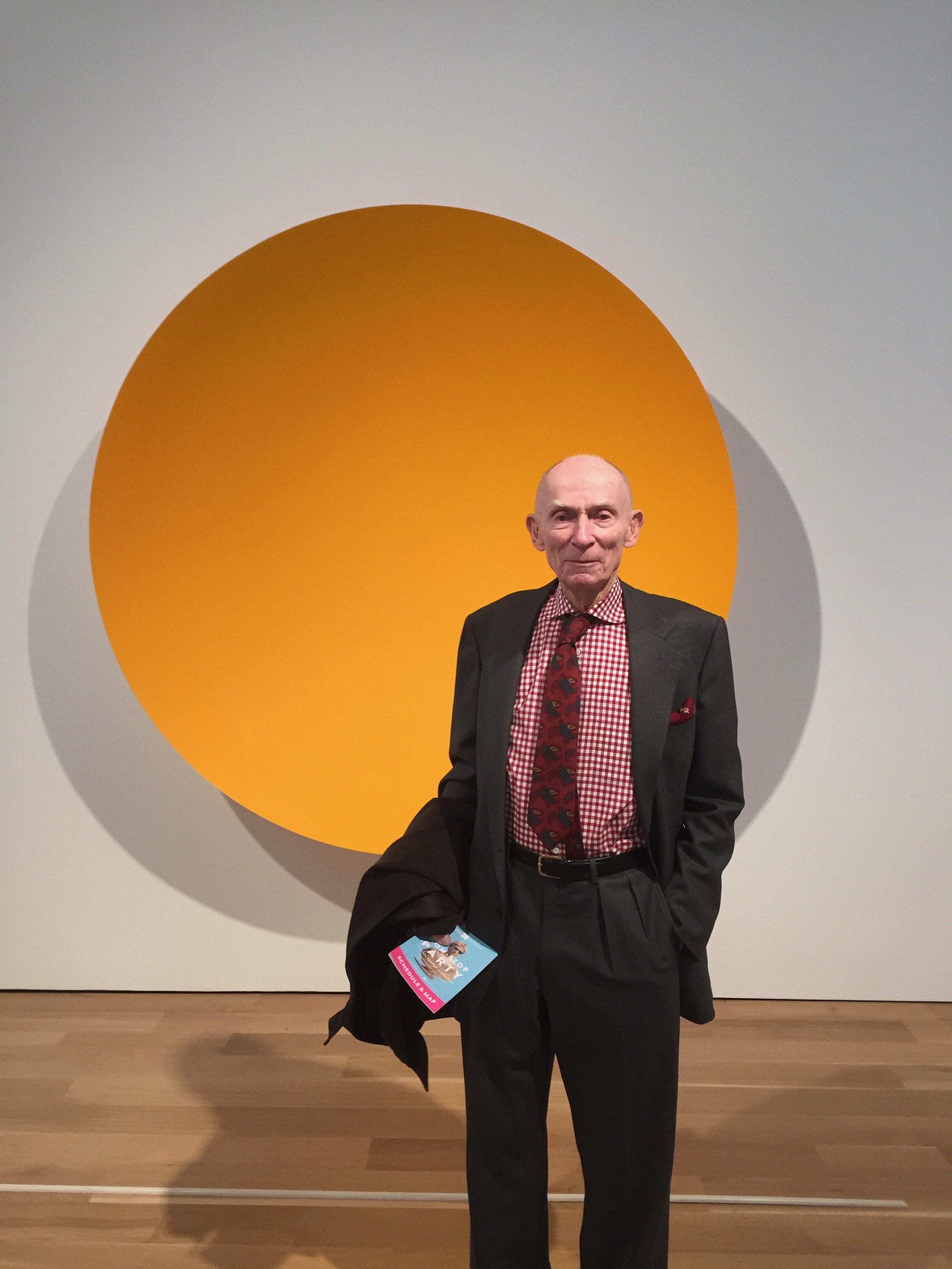 Al Shands, a senior man, is wearing a black suit jacket and matching pants, a gingham button up, and a maroon neck tie, and holds a coat in one arm with a small blue exhibition program, and the other hand in his pocket, looking directly forward in a gallery in front of a large, circular painting of a solid field of yellow and orange, that resemble the sun.