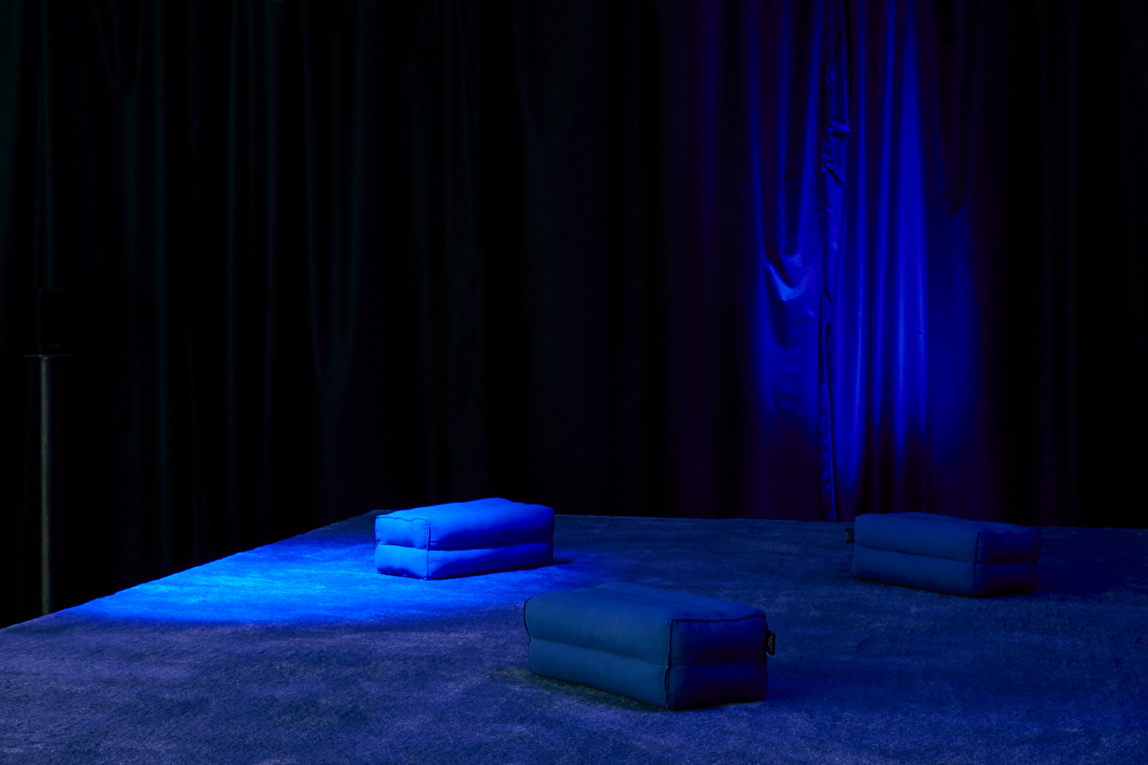 The image shows the sound work Women Sitting at the Machine, Thinking as installed at CCA Derry-Londonderry in 2018, the bottom third of the image shows the surface of a blue carpeted platform, with three blue bolster pillows resting on it. A wide black curtain hands in the background. It is dimly lit, with a diffused spotlight shining from the right onto the surface of the platform.