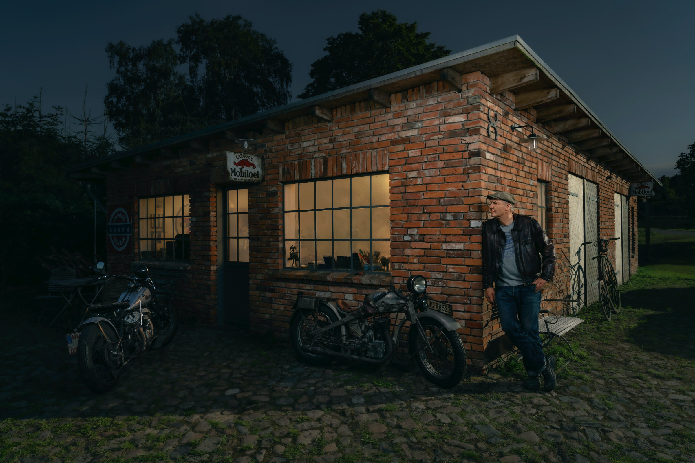 Environmental Portrait aus der Serie The Workshop mit einem Opel-Motorrad. Backsteinwerkstatt im Grünen in dunkler Atmosphaere. Vor der Werkstatt sieht man zwei Oltimermotorraeder und einen Mann.