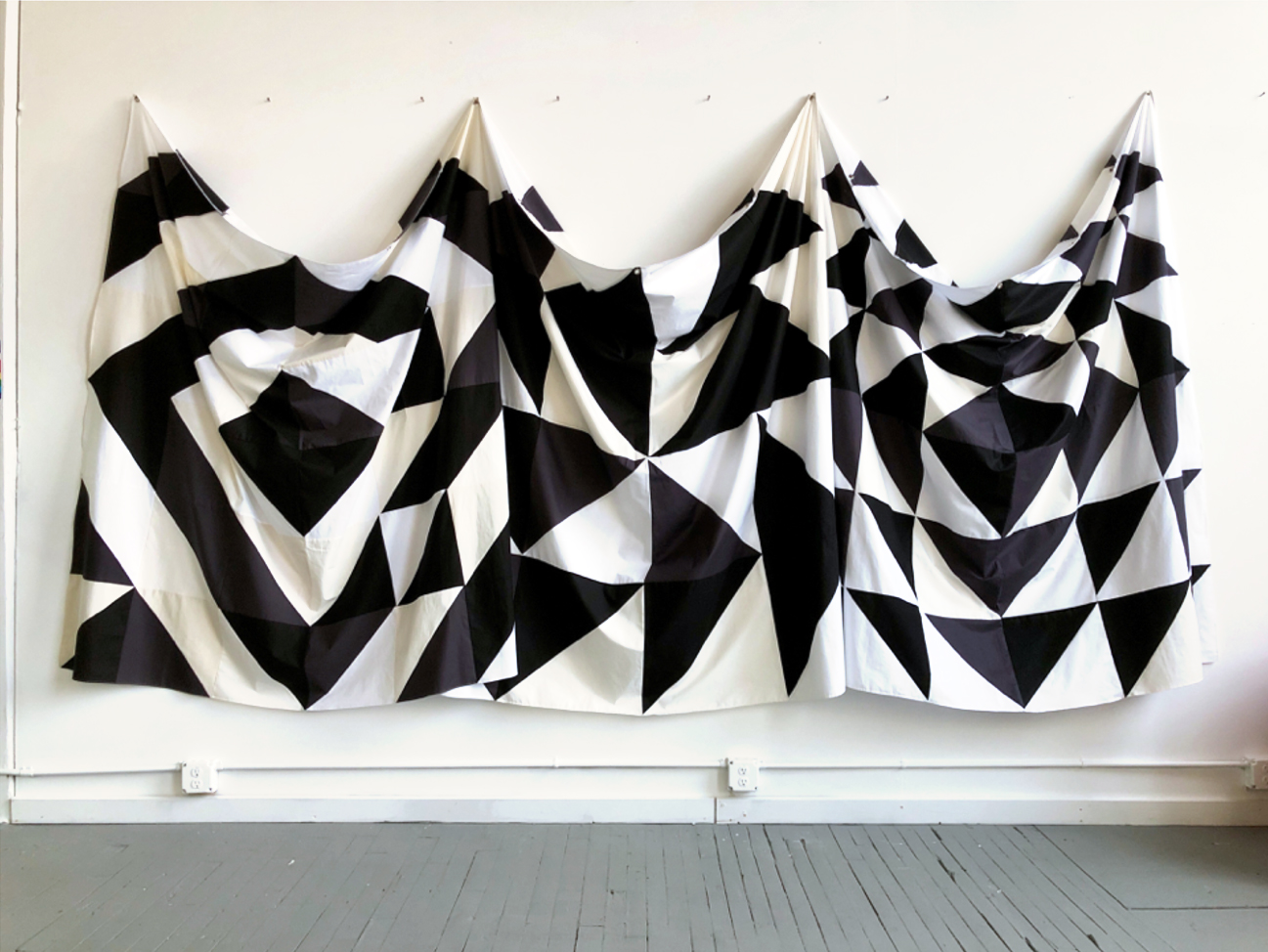 A long, rectangular piece of canvas, like a banner or theatre curtain, hangs on a white wall and drapes from 4 hooks just above a grey painted hardwood floors. The canvas is painted with a black and white geometric design mostly comprised of diamonds and triangles arranges is various patterns.