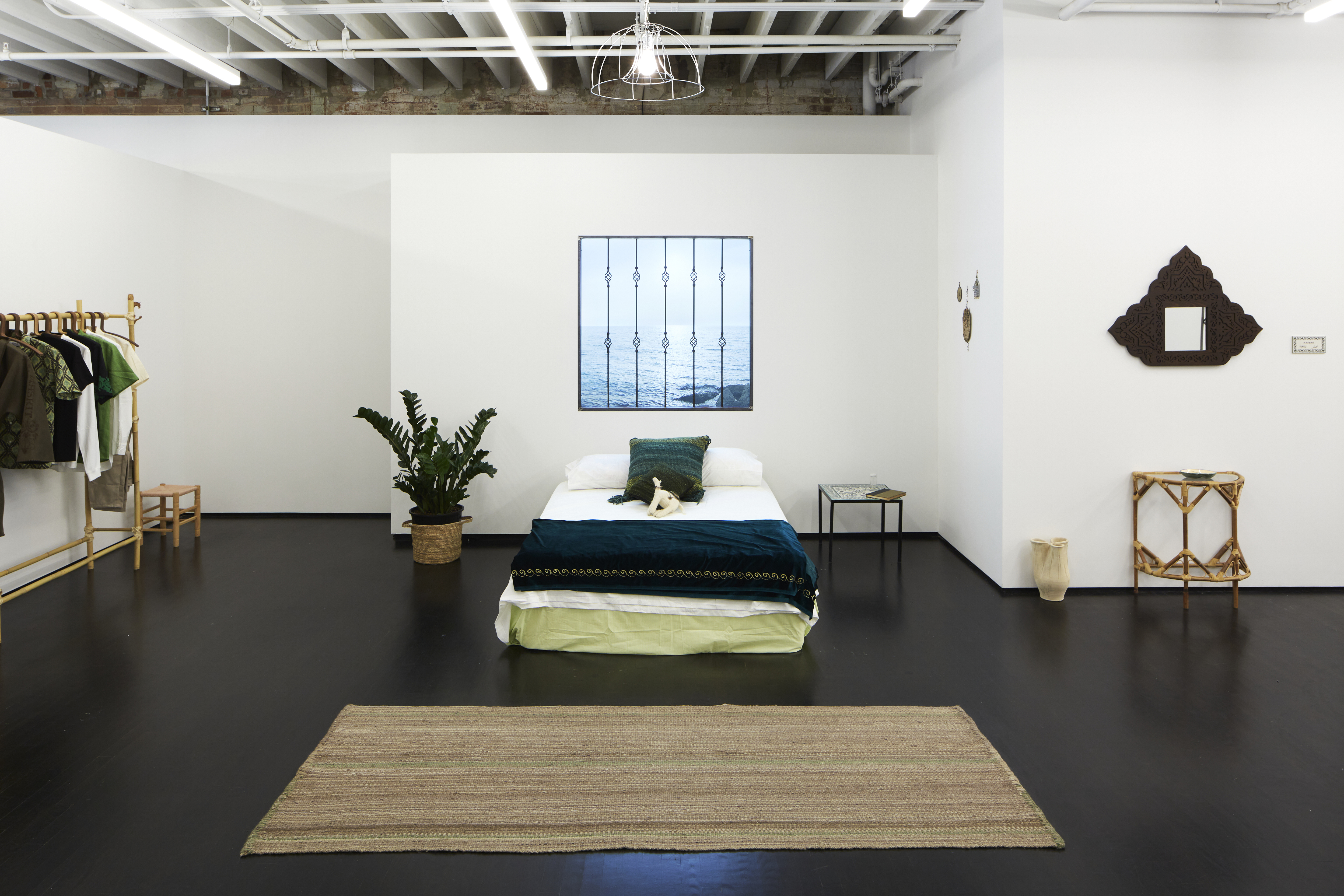 Another view of the home installation, this time with another lightbox in the middle, behind a bed with a green blanket and green pillow. Off to the left is a wooden rack for hanging clothes hooks, which has a set of 4-5 garments on it. Other sparse home furnishing are on the walls and floors.