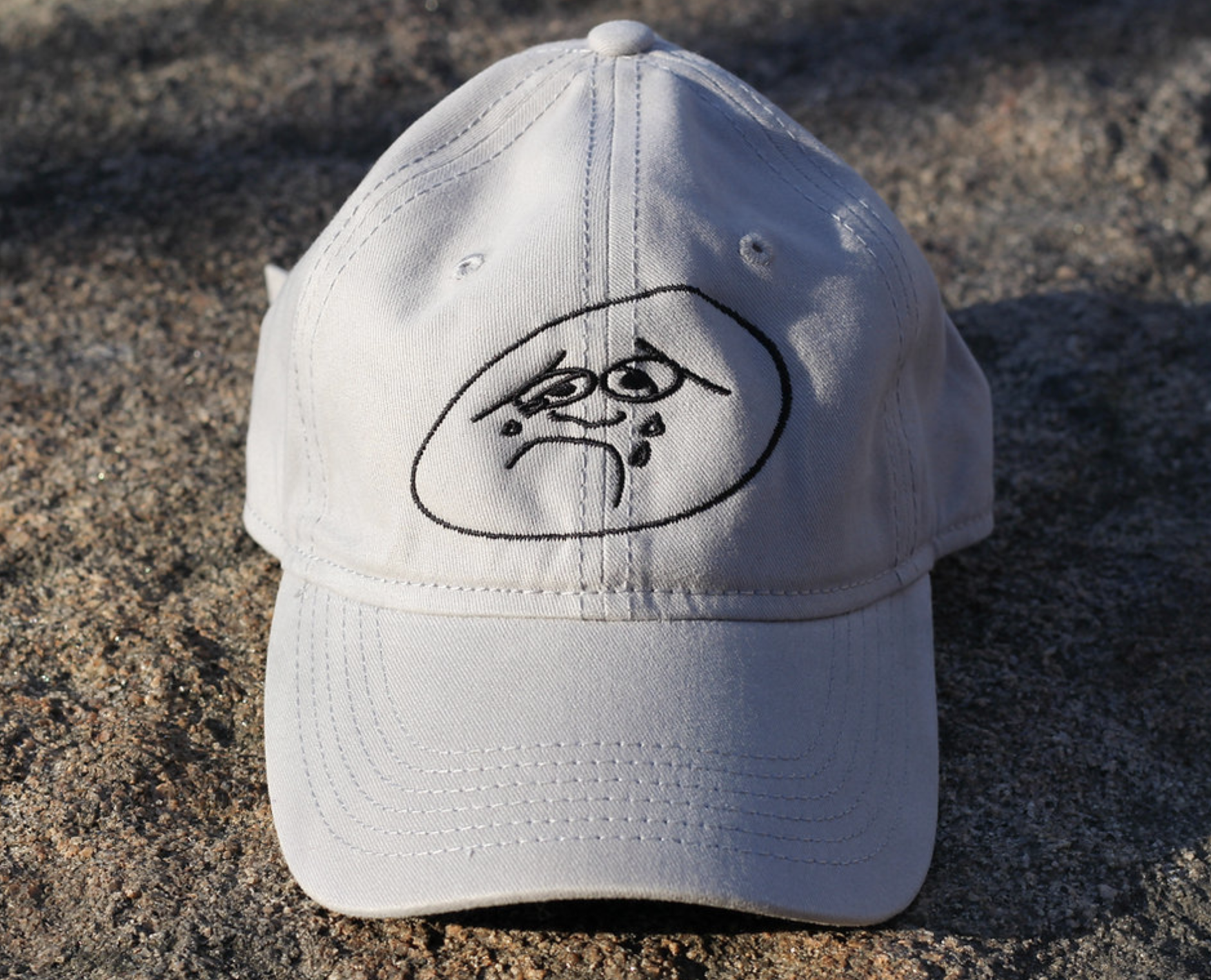 A white baseball cap is centered on a mottled sandy rock surface. A stylized black line drawing of a crying emoji-type face sits above the brim.