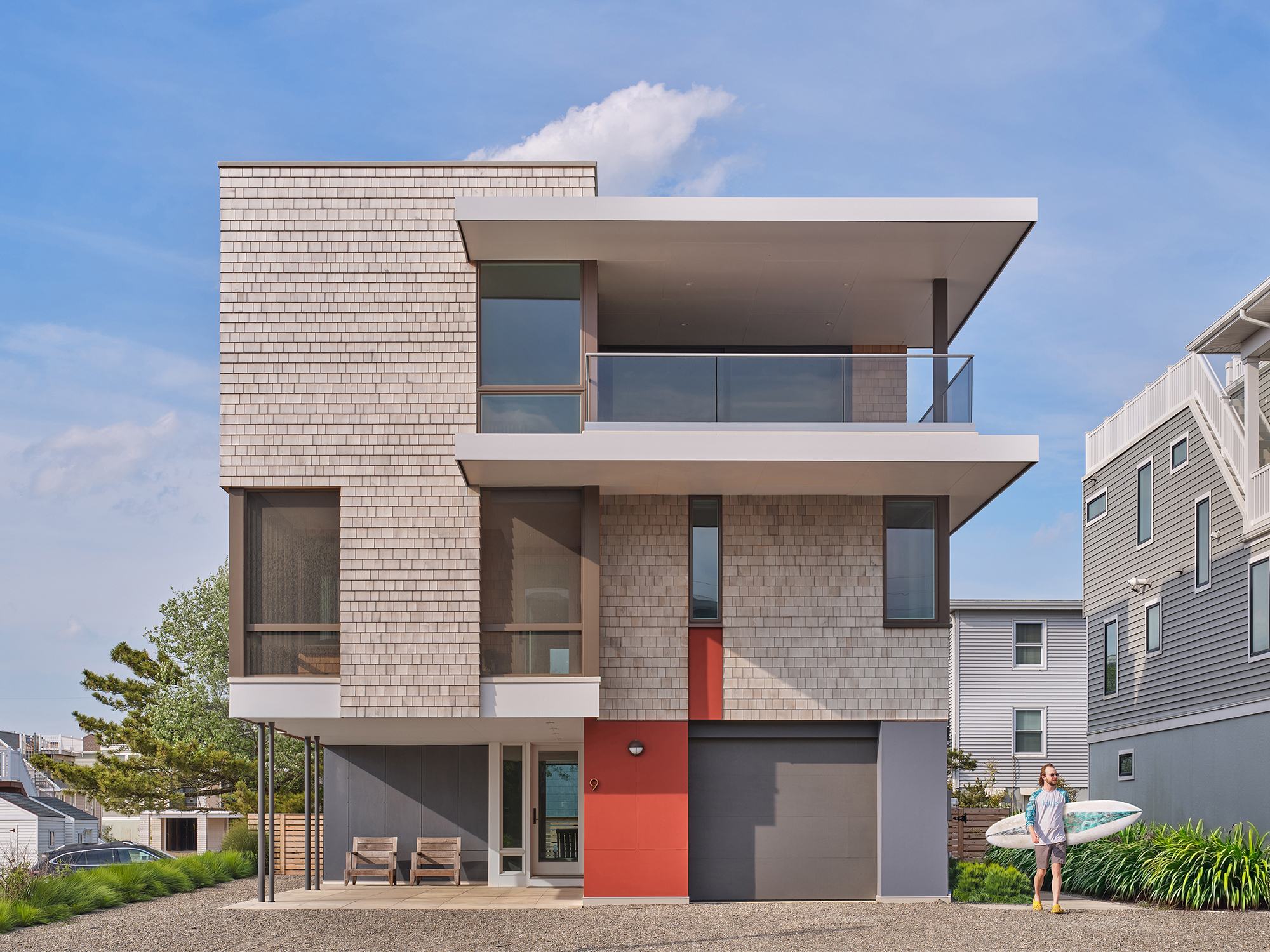 Exterior view of the Gap House South Elevation, featuring cedar shingle siding, a two-car garage, a modern entry with red accent, and a person with a surfboard on the right.