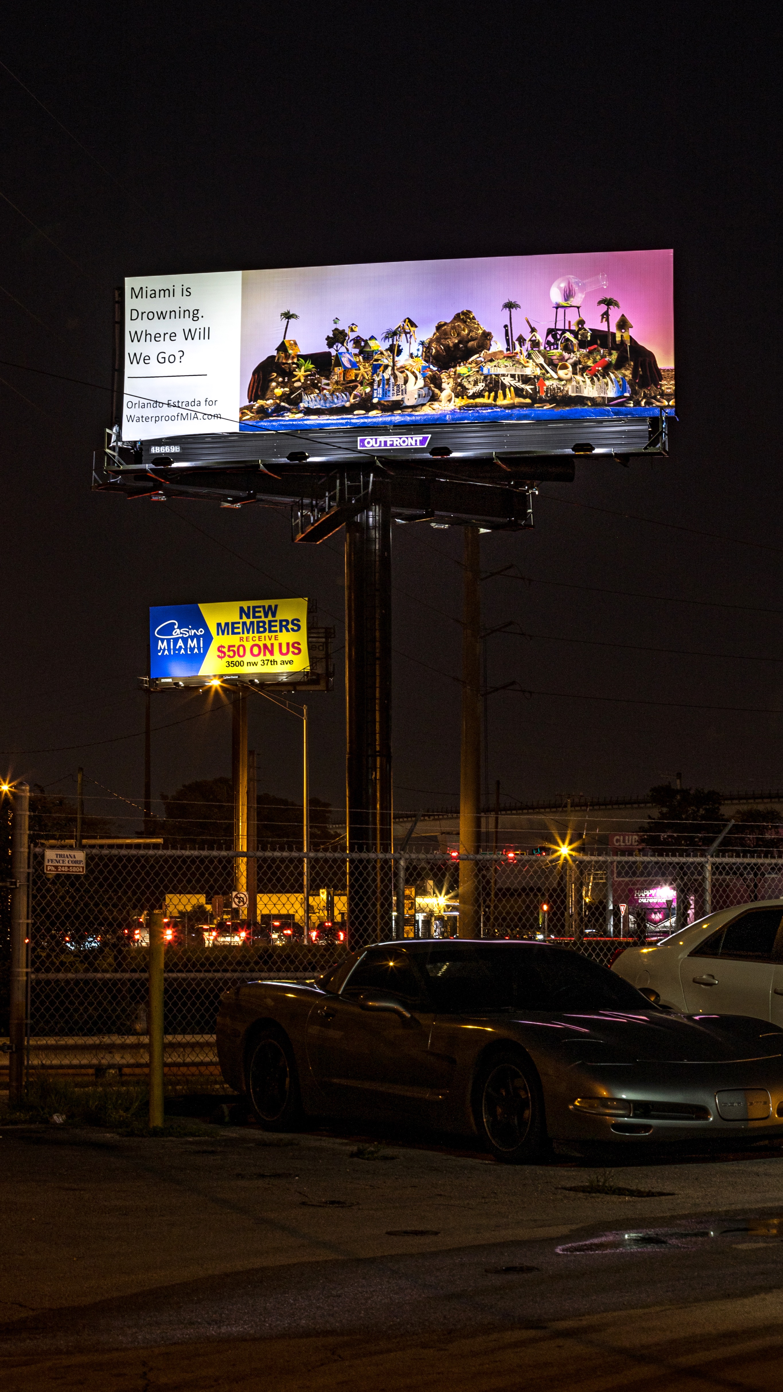 At night, a billboard is displaying an island sunset with a tropical heaven on the left side and a dystopian hell on the right beside a text box that reads in black, “Miami is Drowning. Where Will We Go? Orlando Estrada for WaterproofMIA.com”. A sports car is visibly parked in the foreground.