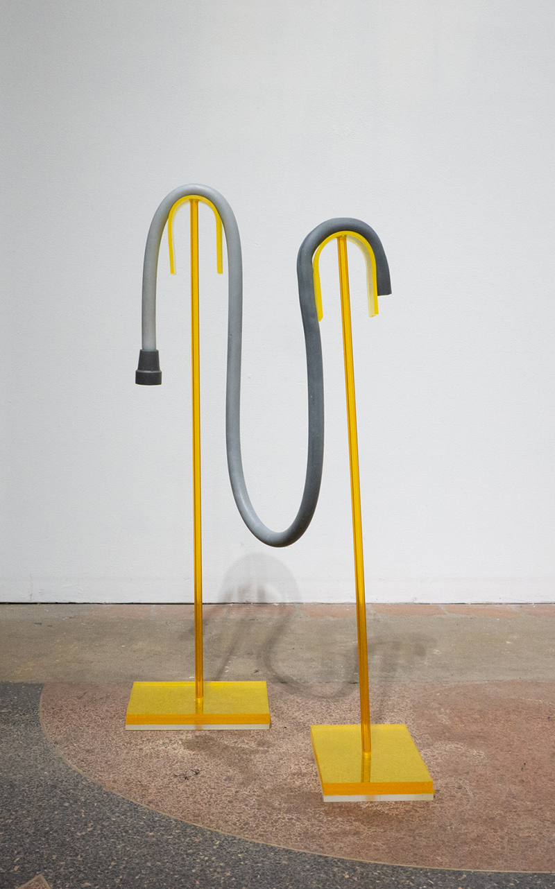 An exaggeratedly long gray rubber cane drapes between two a yellow transparent acrylic stands. The two yellow acrylic stands are each made up of upside down u shapes for the rubber cane’s handle and center to rest on, a vertical acrylic rod and a rectangular platform, all of which are a bright translucent yellow. The cane’s form distorts over these two stands.