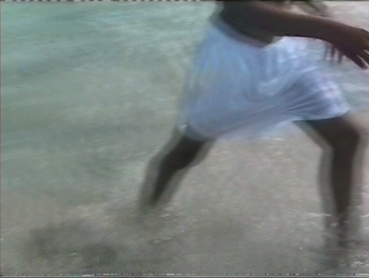 A still from a digitised VHS Tape. A dark-skinned Black person wearing white shorts wades through the water, and out of frame. The image is cropped so that only their lower body is visible; both legs are slightly blurred, feet lost beneath the water, moving in sync with the Caribbean sea. The water is also a blur, starting as a light blue-green and turning into a warm grey as it reaches the shore. The person’s right arm is raised, frozen in the image because they were swinging their arms. They are walking out of the frame.