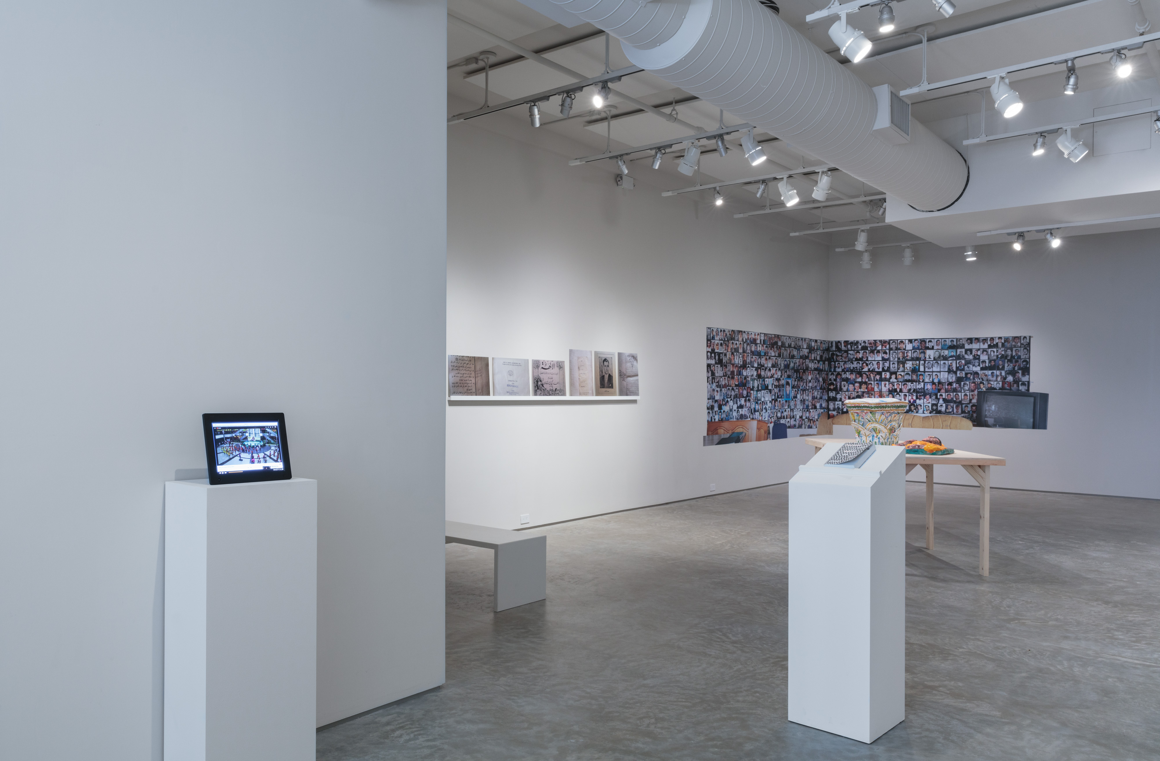 Installation shot, A Void curated by Paul Ramírez Jonas. From left to right: Constanza Ramirez Molano,  Vivificar, 2015. Video documentation, 13 minutes. Emily Jacir, Untitled (fragment from ex libris), 2010-12. 6 c-prints and shelf, 20 5/8 x 115 3/8 1 1/2/52.5 x 293 x 4 cm. Aida Šehović, Family Album (Što Te Nema): Wall 6 and 7, 2018. SAV wallpaper, 126 x 61 & 154 x 61 inches. Michael Rakowitz, May The Obdurate Foe Not Be In Good Health: White limestone Corinthian capital with two rows of acanthileaves, Harem, Byzantine period (5th-6th centuries AD), 2017. Middle eastern packaging and newspapers, glue, 61 x 14 cm. Michael Rakowitz, May The Obdurate Foe Not Be In Good Health (new piece for A Void exhibition), 2018. Middle eastern packaging and newspaper, 24 x 18 x 6 inches. Jonathan Safran Foer,  Tree of Codes, 2010. Paperback, 5.5 x 1 x 8.8 inches. Credit: Stan Narten.