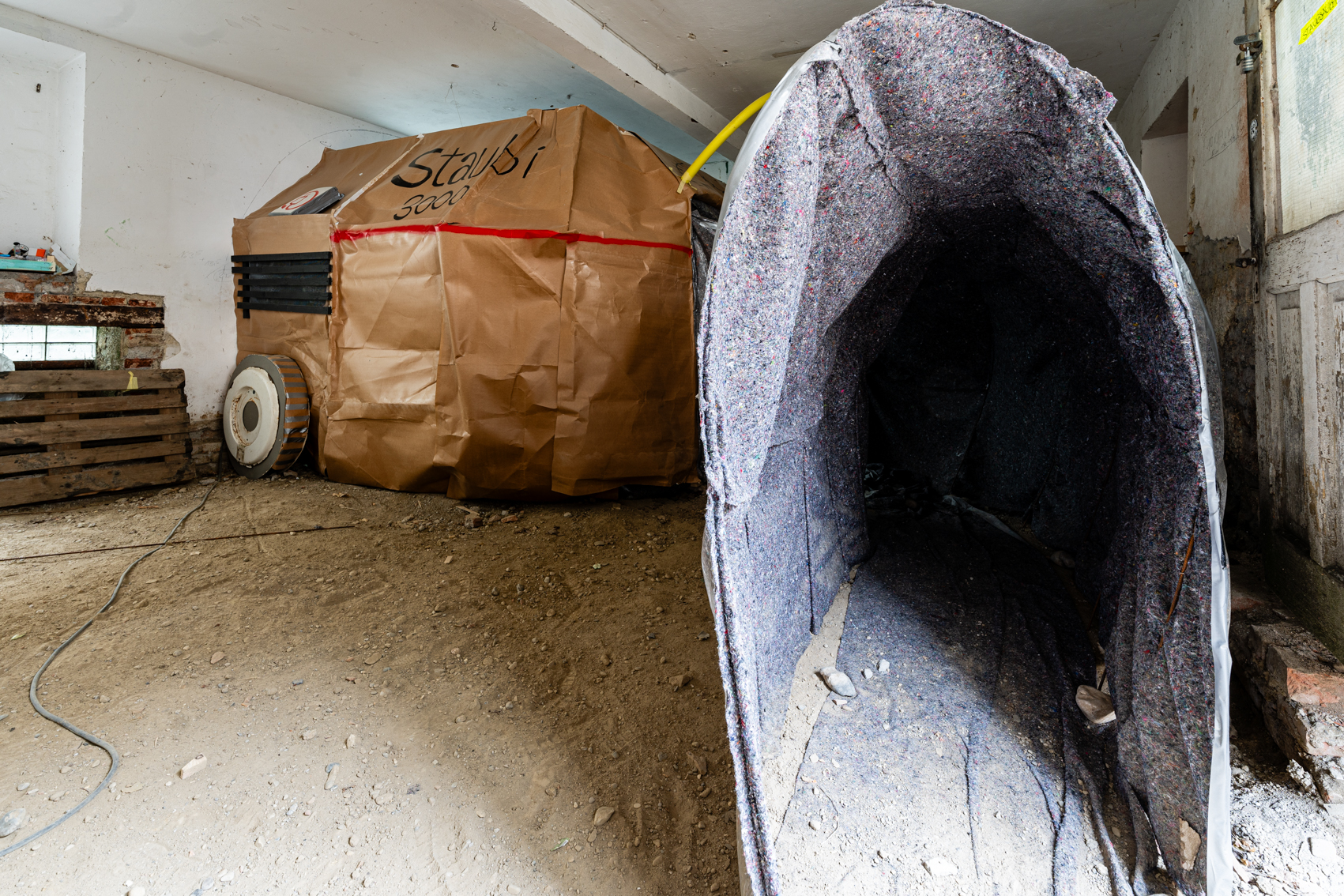 Bild 1 Staubsauber: Vorne ein begehbarer Tunnel aus grauem Teppichflie&szlig;. Im hinteren Teil des Raumes aus Pappkarton ein eigener abgetrennter circa 2 Meter hoher Raum im Raum auf dem Staubi 3000 steht. Dieser ist durch den Tunnel zu erreichen.  