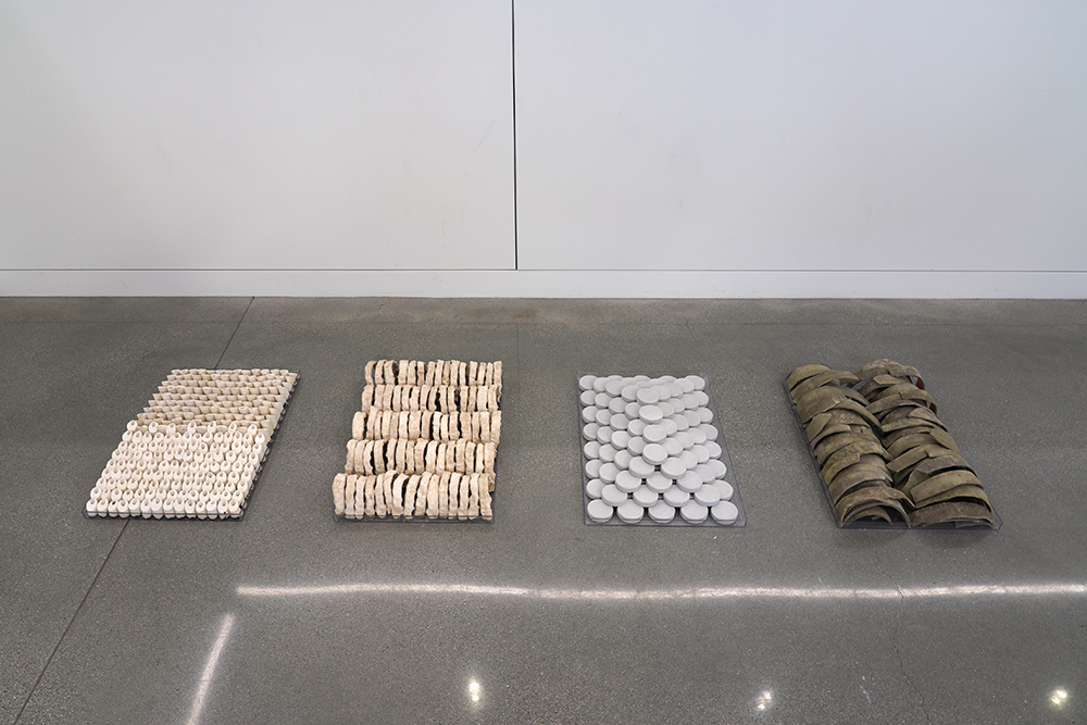 Four trays each filled with multiples of a sculptural piece, installed on the floor.