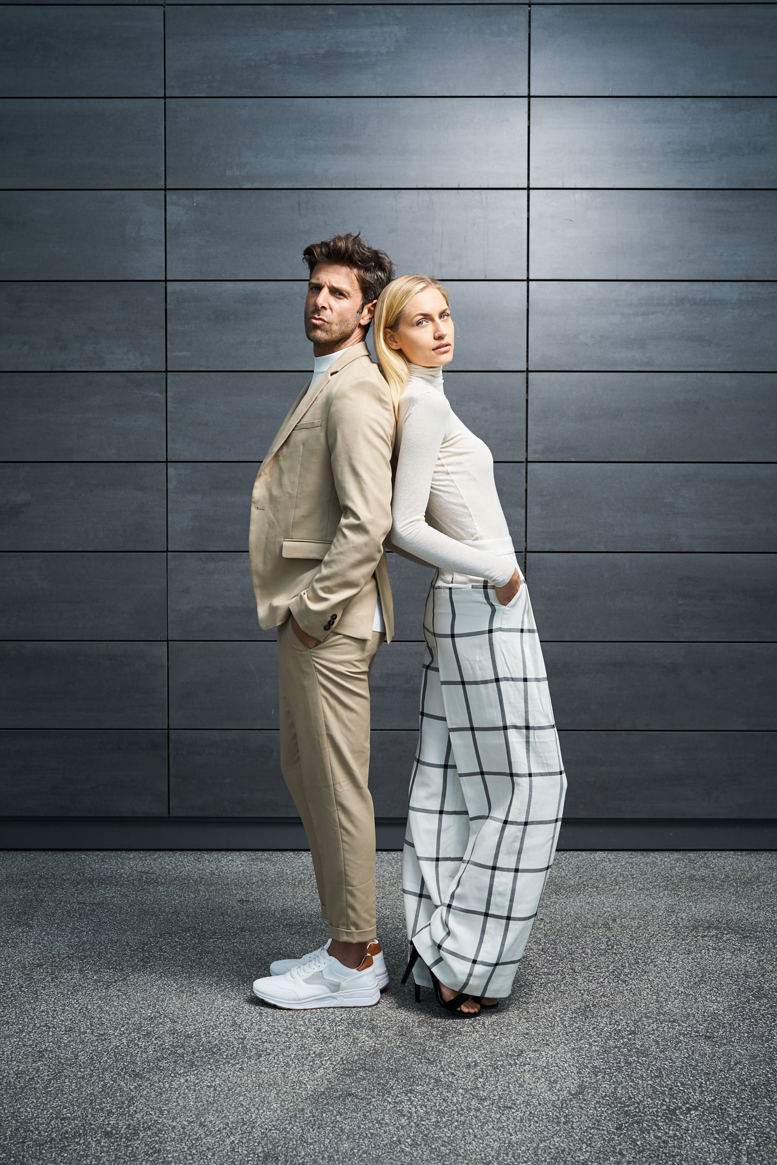 Ein hell gekleidetes Paar steht vor einem grau gekachelten Hintergrund.