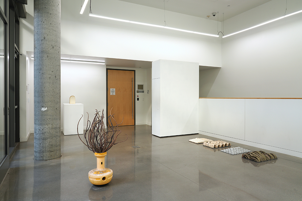 Three sculptures (one standing on the floor, one on a pedestal, and a group of multiples of four shapes, installed on the floor) in a gallery.