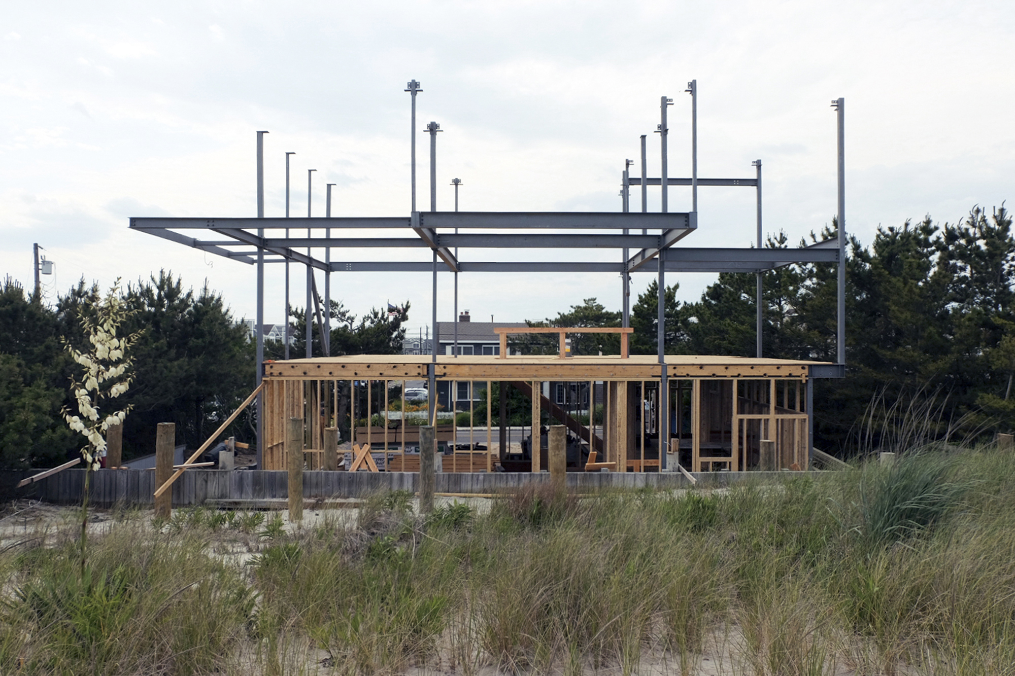Photograph of the Beach Roost residence during the structural erection phase, showing the wood framing of the first floor and the exposed steel frame structure of the second floor against the dune landscape.