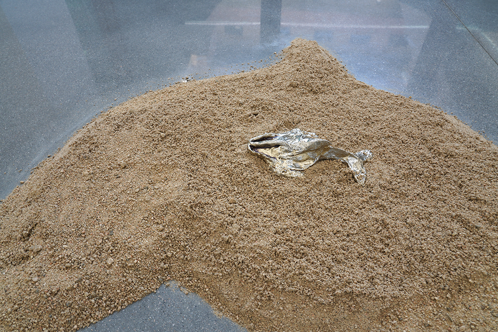 A sculpture in the form of a whale installed on the sand, on the floor.