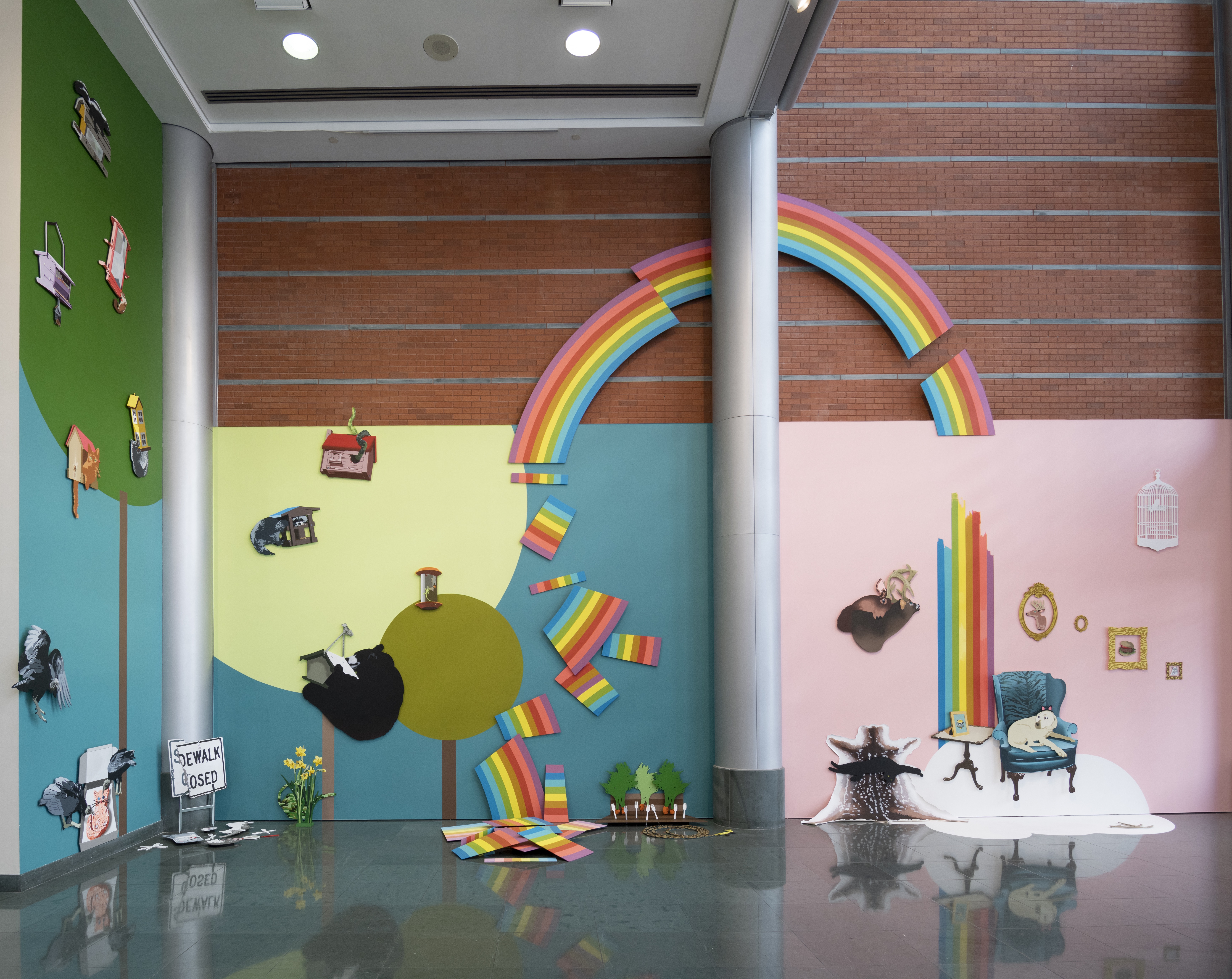 Wide angle view of a colorful art installation that has many differently painted background walls behind flattened painted images of things like a broken up rainbow, a surreal living room scene, trees and a sky.