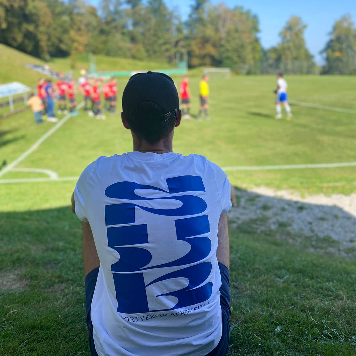 Young man sitting aside the pitch, wearing a white merch shirt of the club