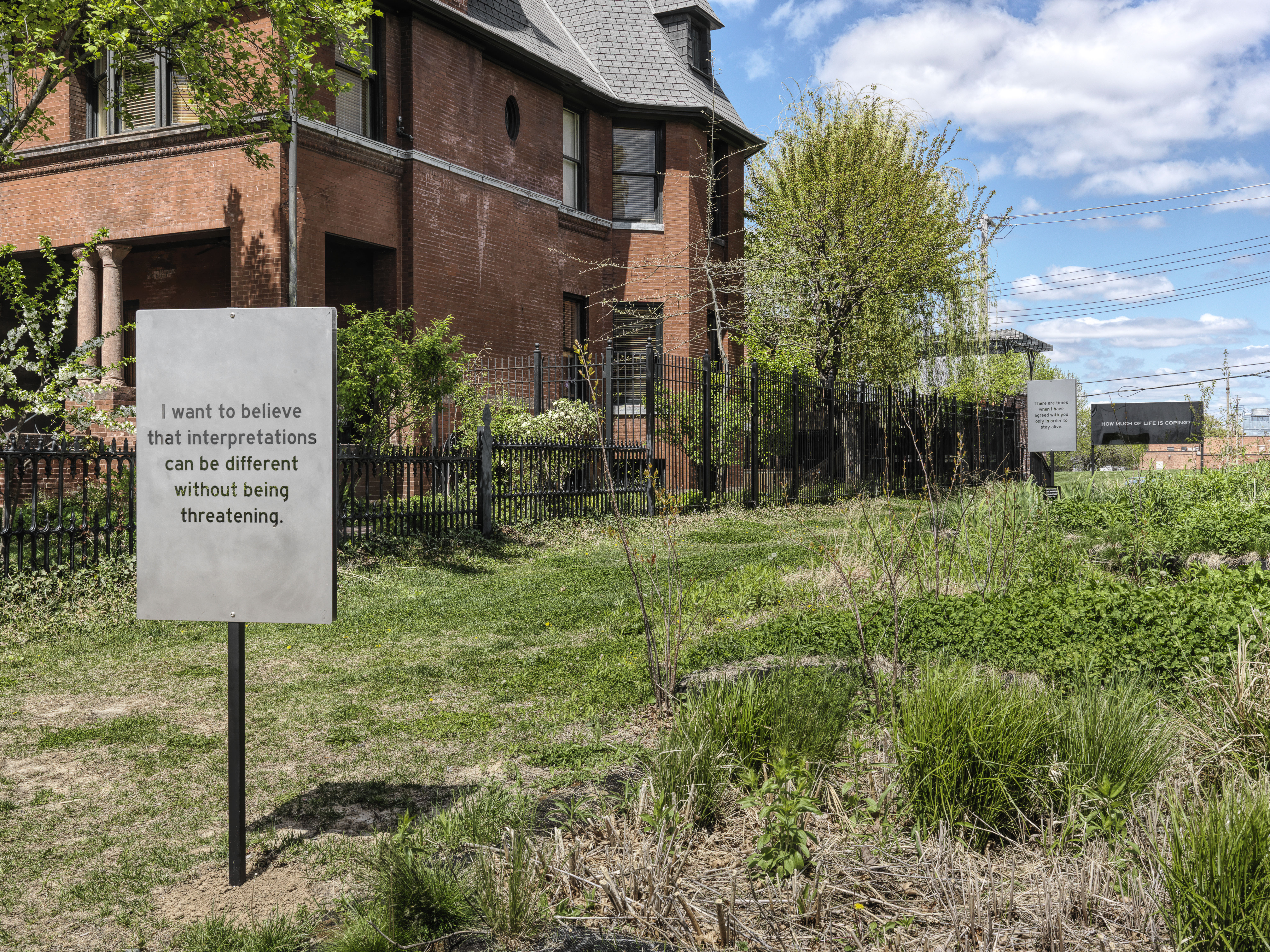 In an empty grassy side lot of a two story residential brick house on a cloudy blue sky day are three signs aligned in a row the length of the property. The nearest, a grey rectangular sign that is taller than it is wide, with black lettering reads: “I want to believe that interpretations can be different without being threatening.”