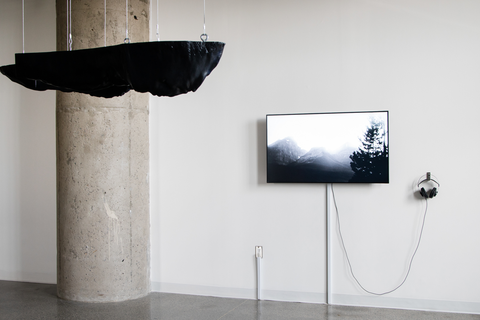 Closer view of the black three-dimensional sculpture by Polymetis. Its top surface is straight and smooth, but in contrast the rest of the sculpture is organic and reminiscent of landscape. Behind it, the black-and-white video of work by Kolcze depicts mountains and trees.