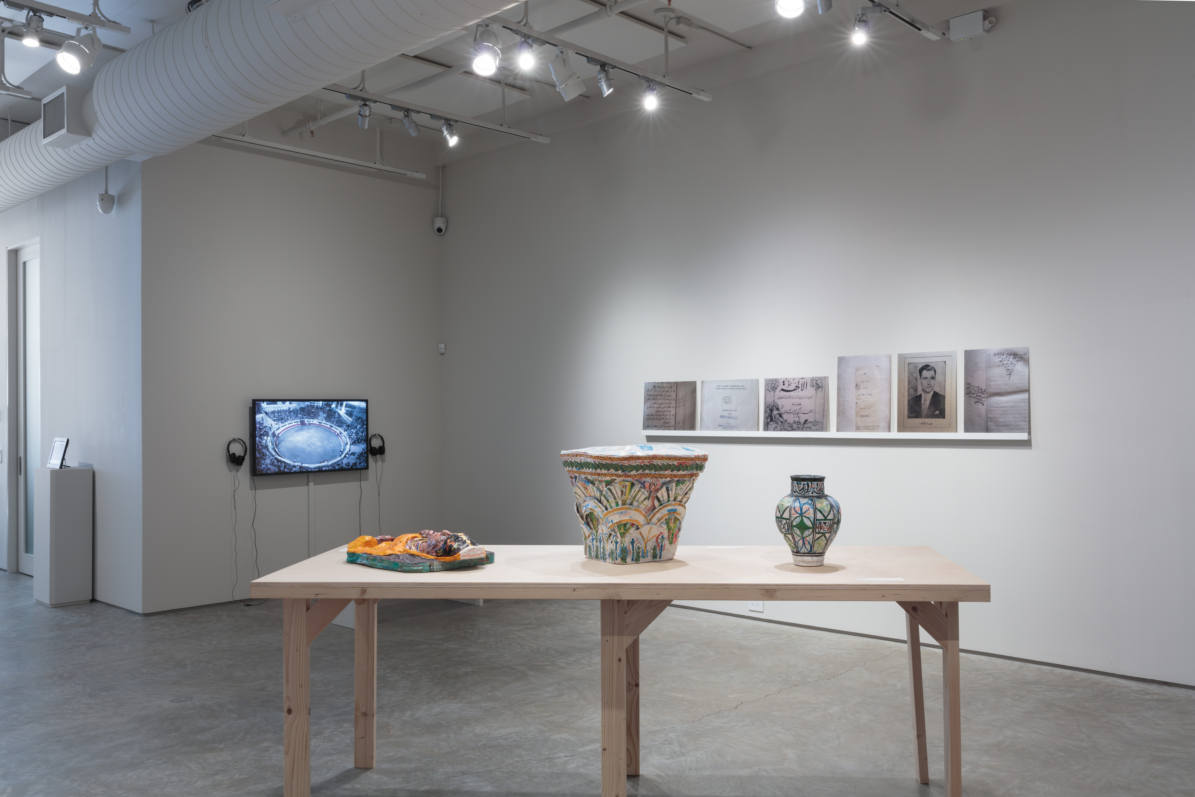 Installation shot, A Void curated by Paul Ramírez Jonas. From left to right: Suzanne Lacy, De tu puño y letra, 2015. Video, 13 minutes. Michael Rakowitz, May The Obdurate Foe Not Be In Good Health (new piece for A Void exhibition), 2018. Middle eastern packaging and newspaper, 24 x 18 x 6 inches. Michael Rakowitz, May The Obdurate Foe Not Be In Good Health: White limestone Corinthian capital with two rows of acanthileaves, Harem, Byzantine period (5th-6th centuries AD), 2017. Middle eastern packaging and newspapers, glue, 61 x 14 cm. Michael Rakowitz, May The Obdurate Foe Not Be In Good Health: Glazed polychrome ceramic vase, 2017. Middle eastern packaging and newspapers, glue, 13.1875 x 10.5 inches. Emily Jacir, Untitled (fragment from ex libris), 2010-12. 6 c-prints and shelf, 20 5/8 x 115 3/8 1 1/2/52.5 x 293 x 4 cm. Credit: Stan Narten.