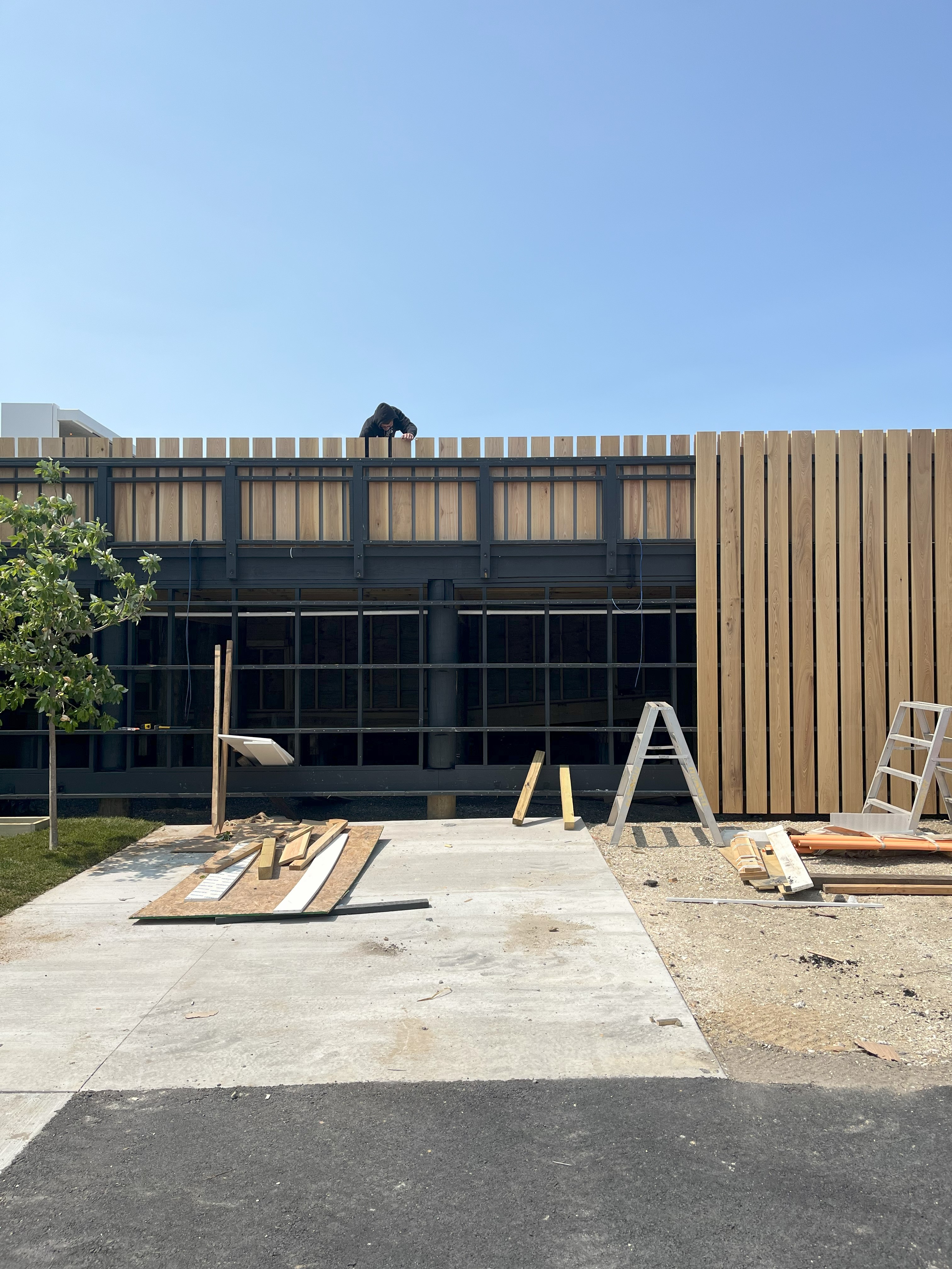 Construction photo showing the ground floor of the Brigantine Marina Paddle Club, featuring dark window openings and vertical wood cladding being installed in the foreground.