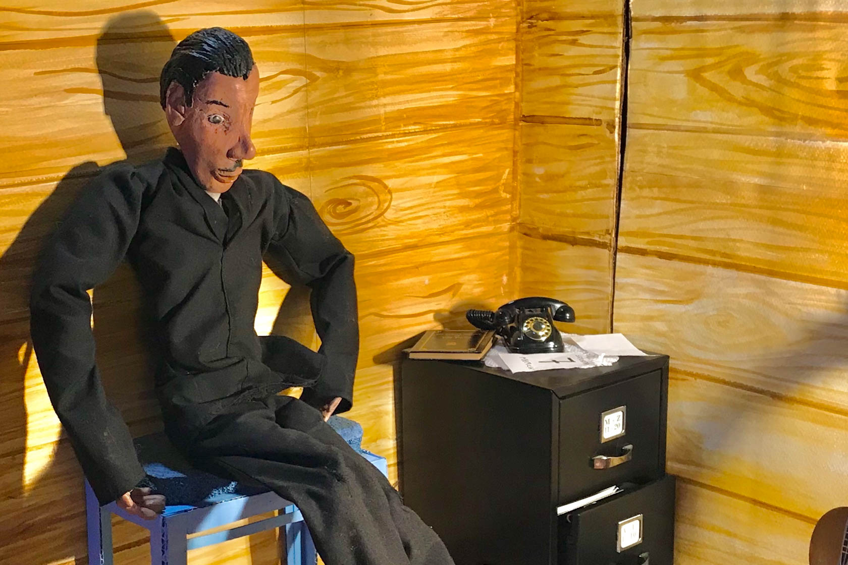 A miniature male puppet sits alone in the corner of a room, back pressed against the wall.