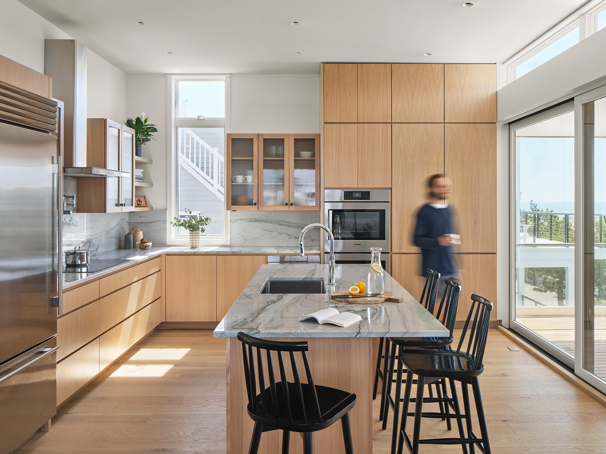 Architectural render of the Gap House kitchen, featuring a central island with seating, light wood custom cabinetry, integrated appliances, and sliding glass doors opening to an ocean-facing deck.