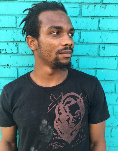 Headshot of Shaun, who is looking off to the right in front of a bright teal-blue painted brick wall. He is wearing a black t shirt with a graphic design over his heart.