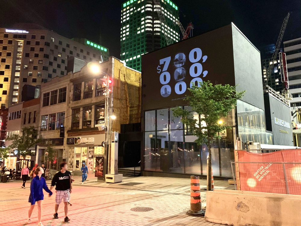 Saint Catherine Street in Montreal at night; the number "$700 000 000" is projected on a condominium sales office across the street. People are walking past and there are skyscrapers in the background.