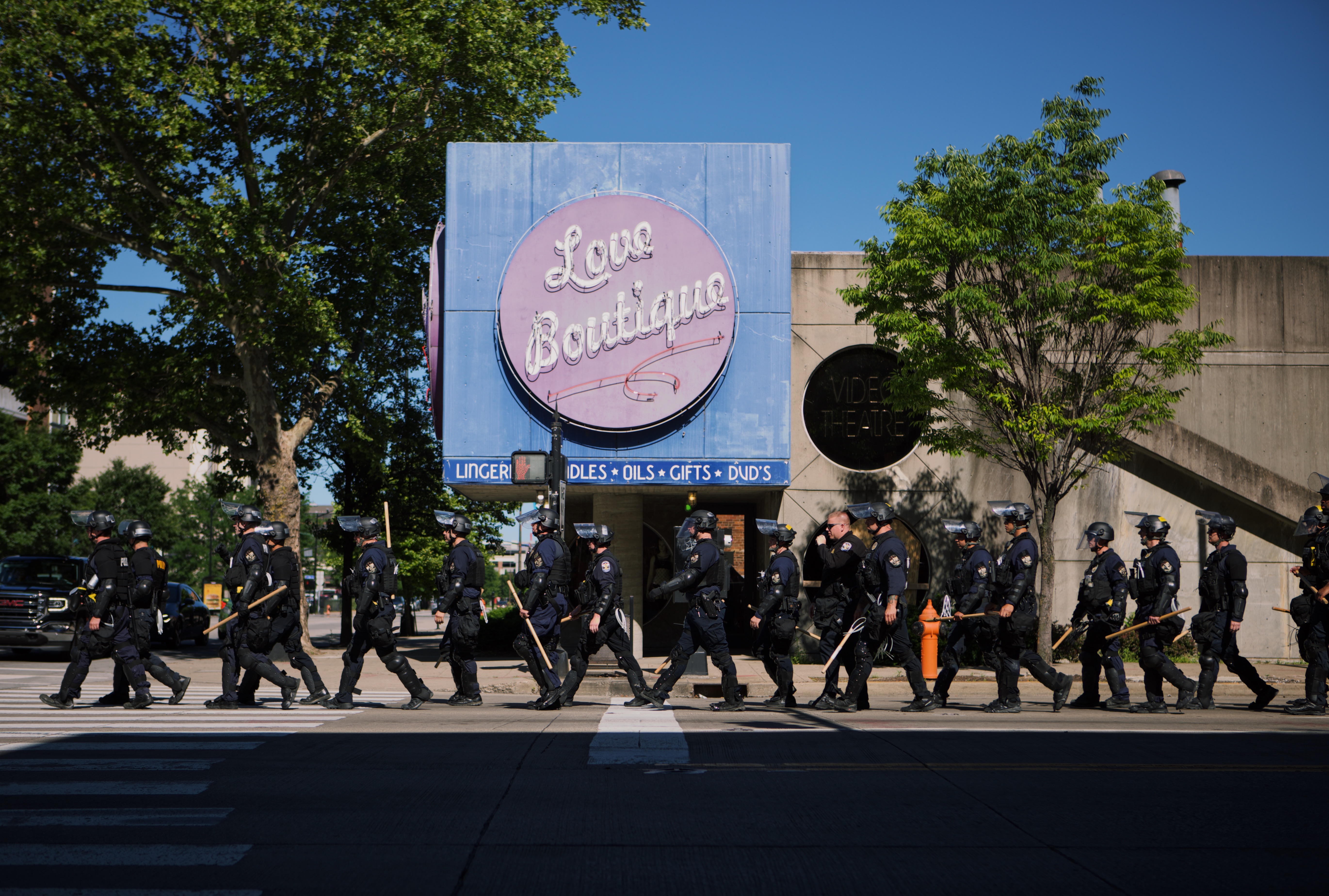 A color photo of several dozen armed police officers in riot gear walk in a line in front of a corner store with a large pink sign that says “Love Boutique” in white script font.