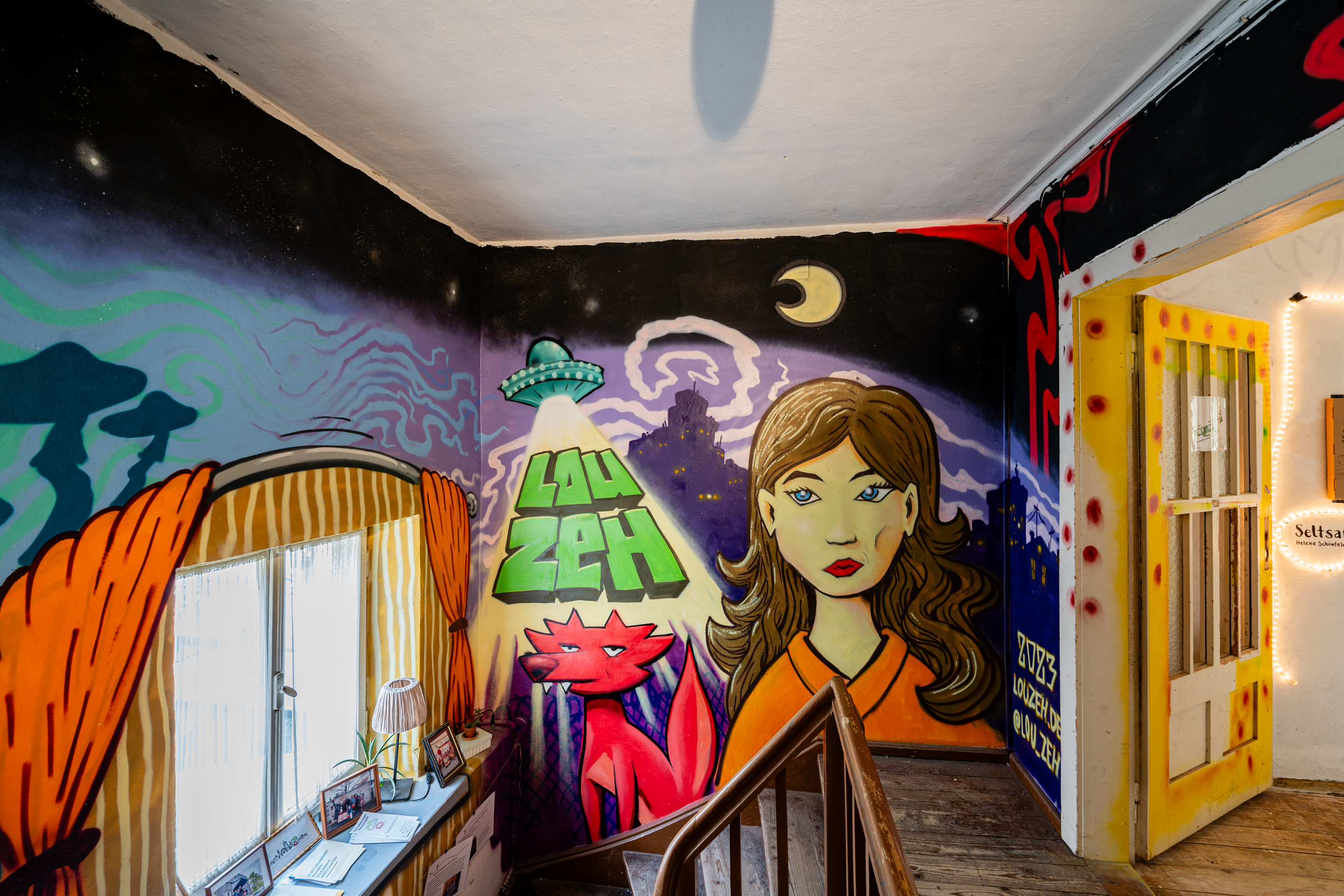 Bild 1 zu Beyond Wildest Dreams: Bunt besprayetes Treppenhaus des obersten Stockwerks. Das Graffiti zeigt einen lilanen von Schwaden &uuml;berzogenen Himmel, im Hintergrund eine Stadt und ein Ufo. In Comic-Stil wurde ein roter Fuchs sowie das Gesicht einer jungen Frau mit braunen Haaren gesprayt. An der linken Wand sind in orange Vorh&auml;nge um das Fenster gemalt worden. 