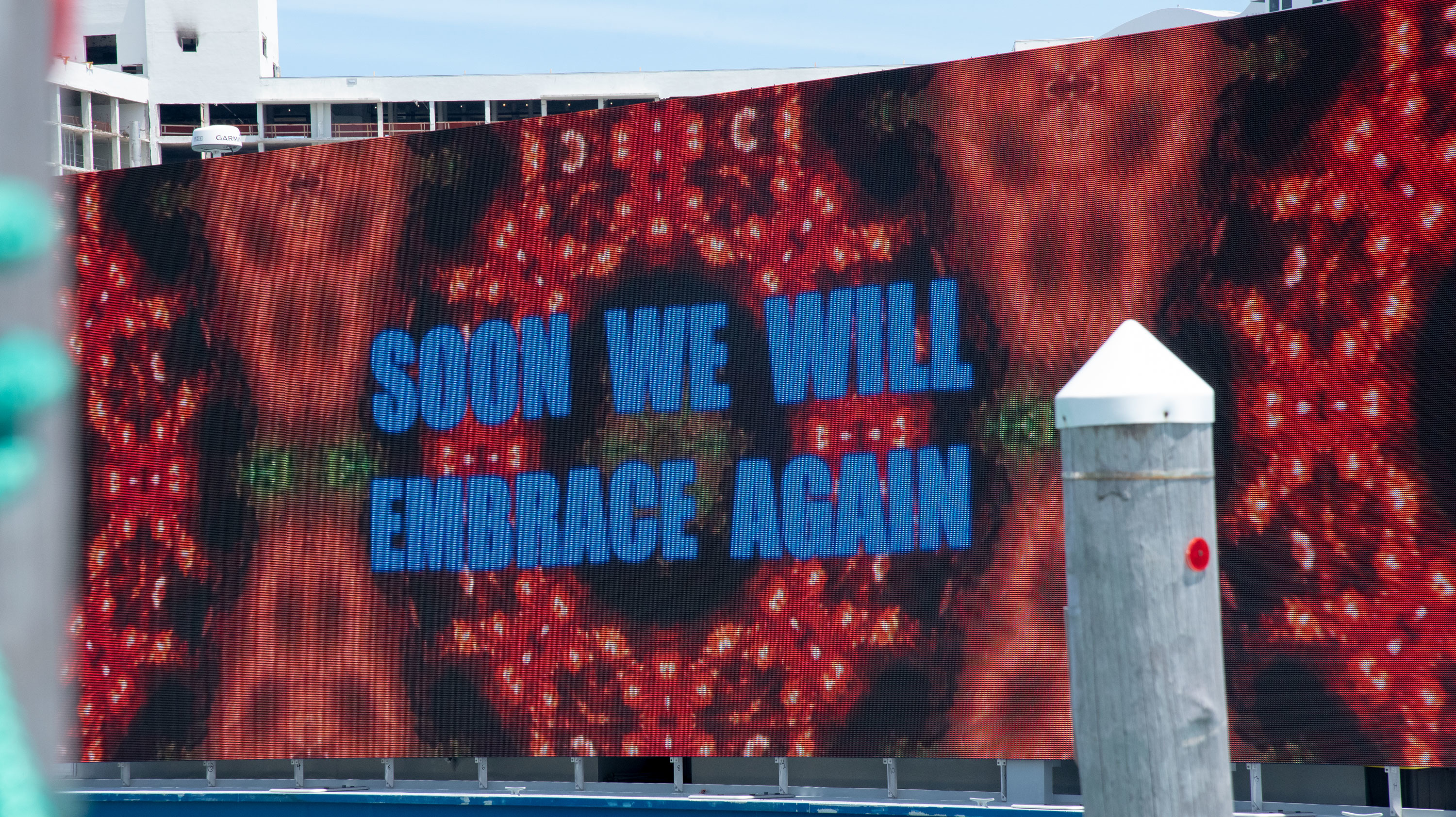 Displayed on a large screen and exhibited by boat in Miami’s Biscayne Bay is Carlos Betancourt’s 2020 video installation.  In large, blue, blocked letters reads the message: “SOON WE WILL EMBRACE AGAIN”.  In the background are large mirroring red kaleidoscope images that have green accents in the center.