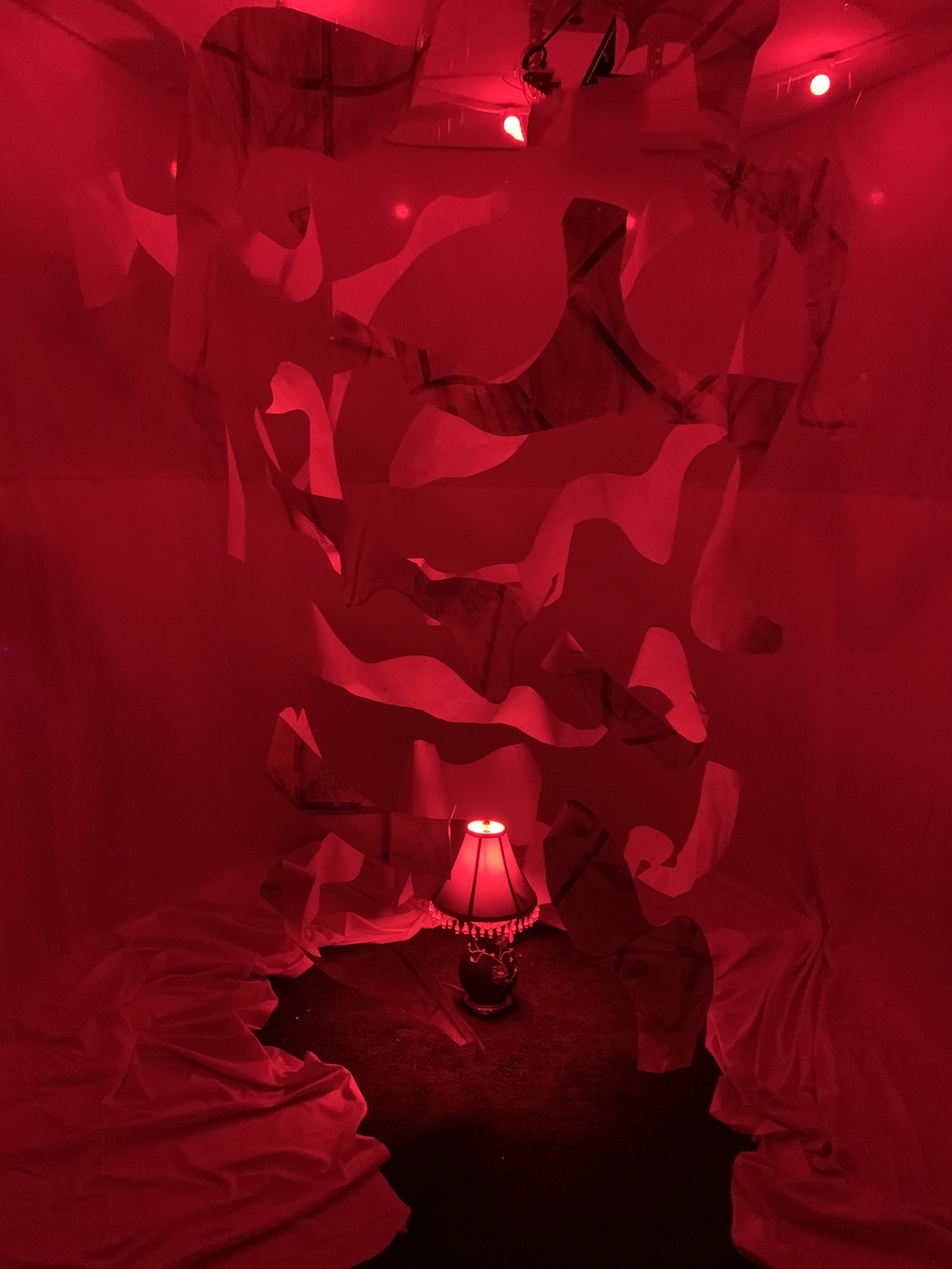 A full view of the sculpture of suspended paper that forms into a hive-like cylinder. Wall to ceiling neutral fabric circles the sculpture to contain and illuminate the space while above a circle of red lights and a lamp under neath illuminates everything in red