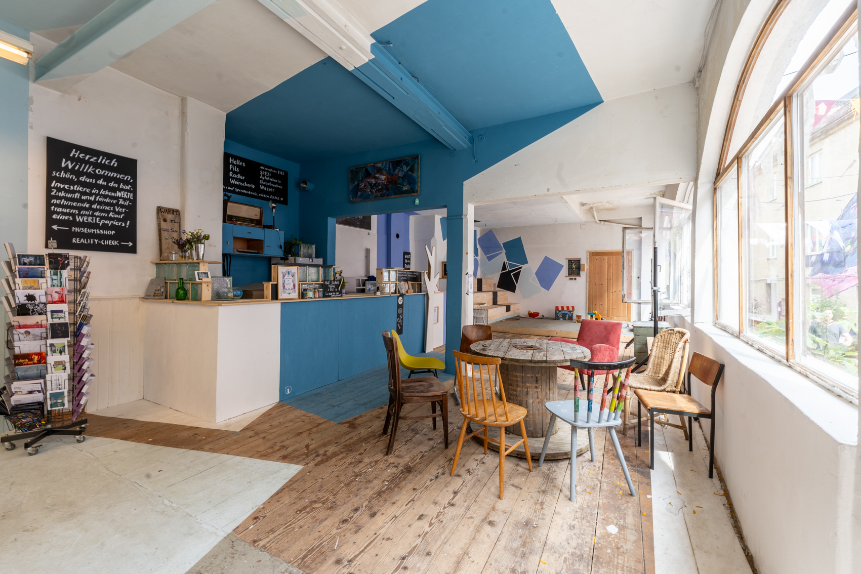 Bild 1 zu Cafe Eder: Blick von der Eingangst&uuml;r in das gro&szlig;e helle Cafe mit blauen Elementen. Die rechte Seite des Raumes zeigt zum Innenhof und ist ges&auml;umt mit einer Fensterfront. Im linken Bereich des Raumes befindet sich die Bar, rechts davon steht ein runder Tisch mit verschiedenen St&uuml;hlen.
