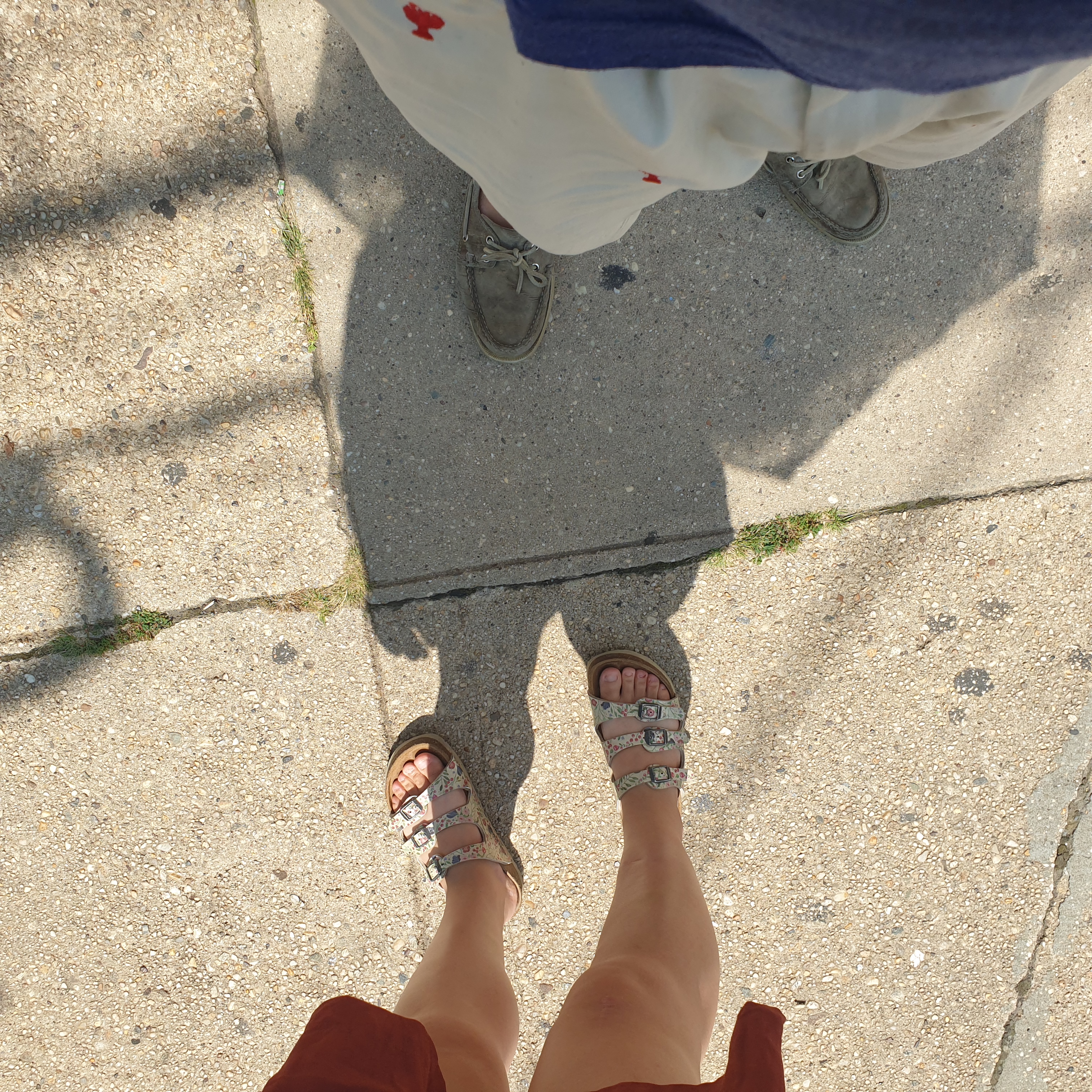 On the right a woman's feet in sparkling sandals, on the left a man's feet in dingy grey boat shoes. The woman's shadow, which includes the outline of her parasol, shades the man. They stand on sidewalk.