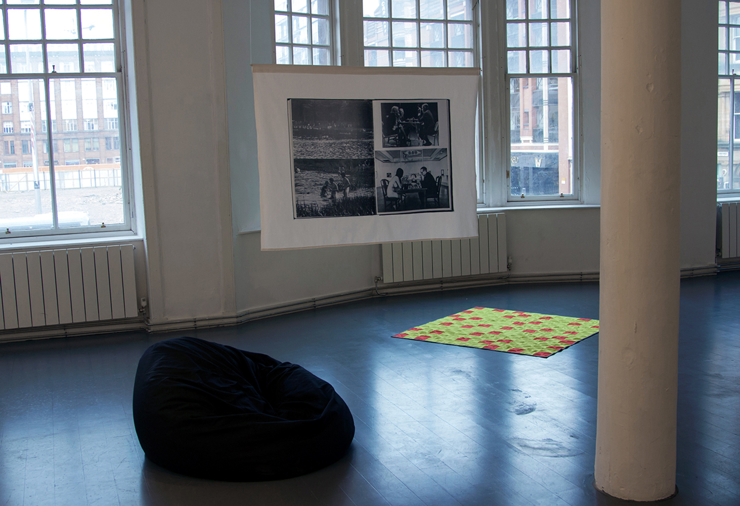 The image shows a mid-shot of the show After Hours, in a white gallery space with a grey floor and windows on the back wall. There is a black bean bag on the floor to the right, and to the left there is a patchwork quilt in high-visibility orange and yellow fabric. A printed fabric banner hangs from the ceiling, showing scanned double page spreads from the catalogue for the landmark 1970 exhibition 'Information' at MOMA, NY.