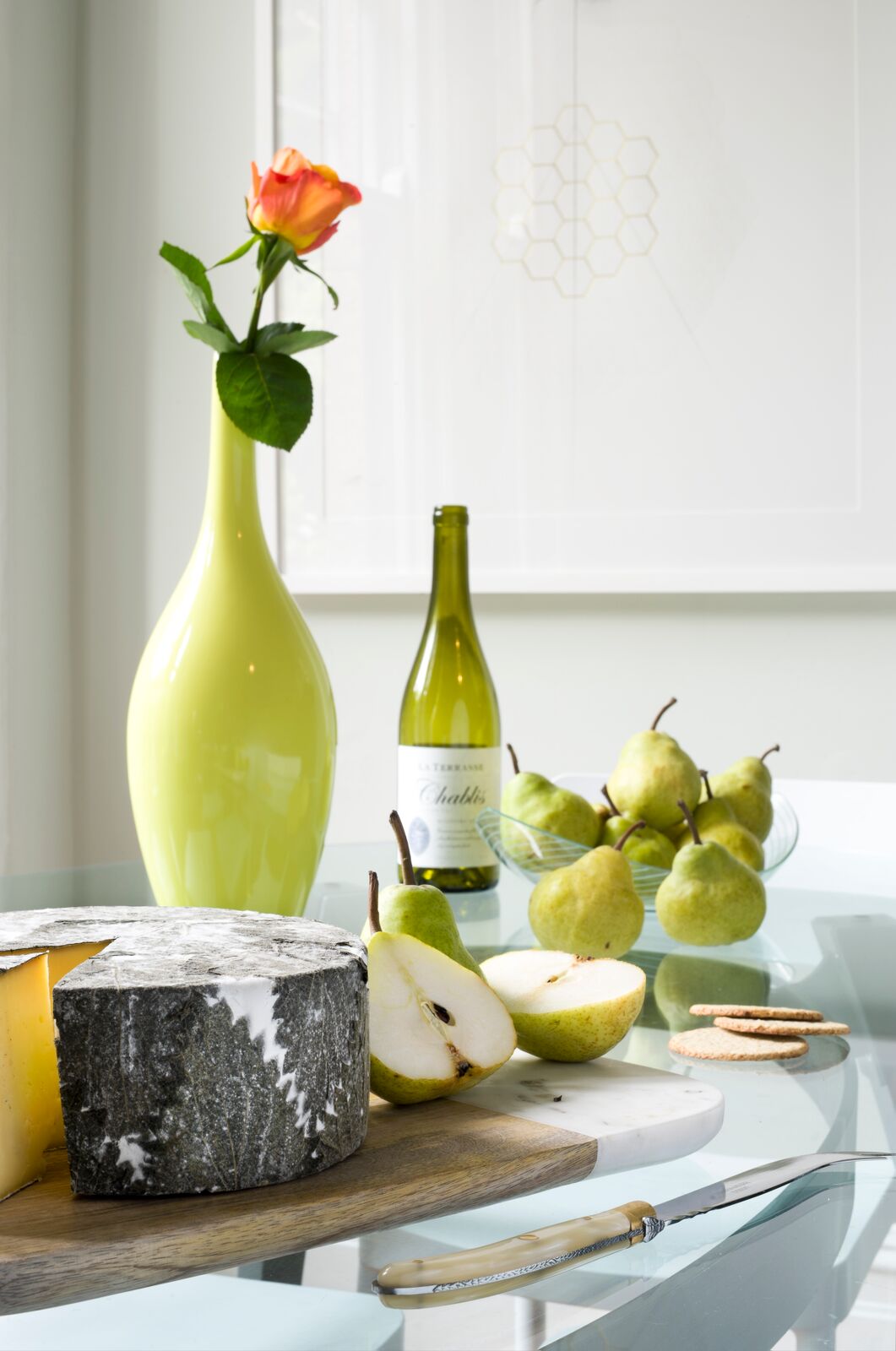 Close-up of cheese board with sliced pears on glass table. Lime green vase with single flower and wine bottle in background.