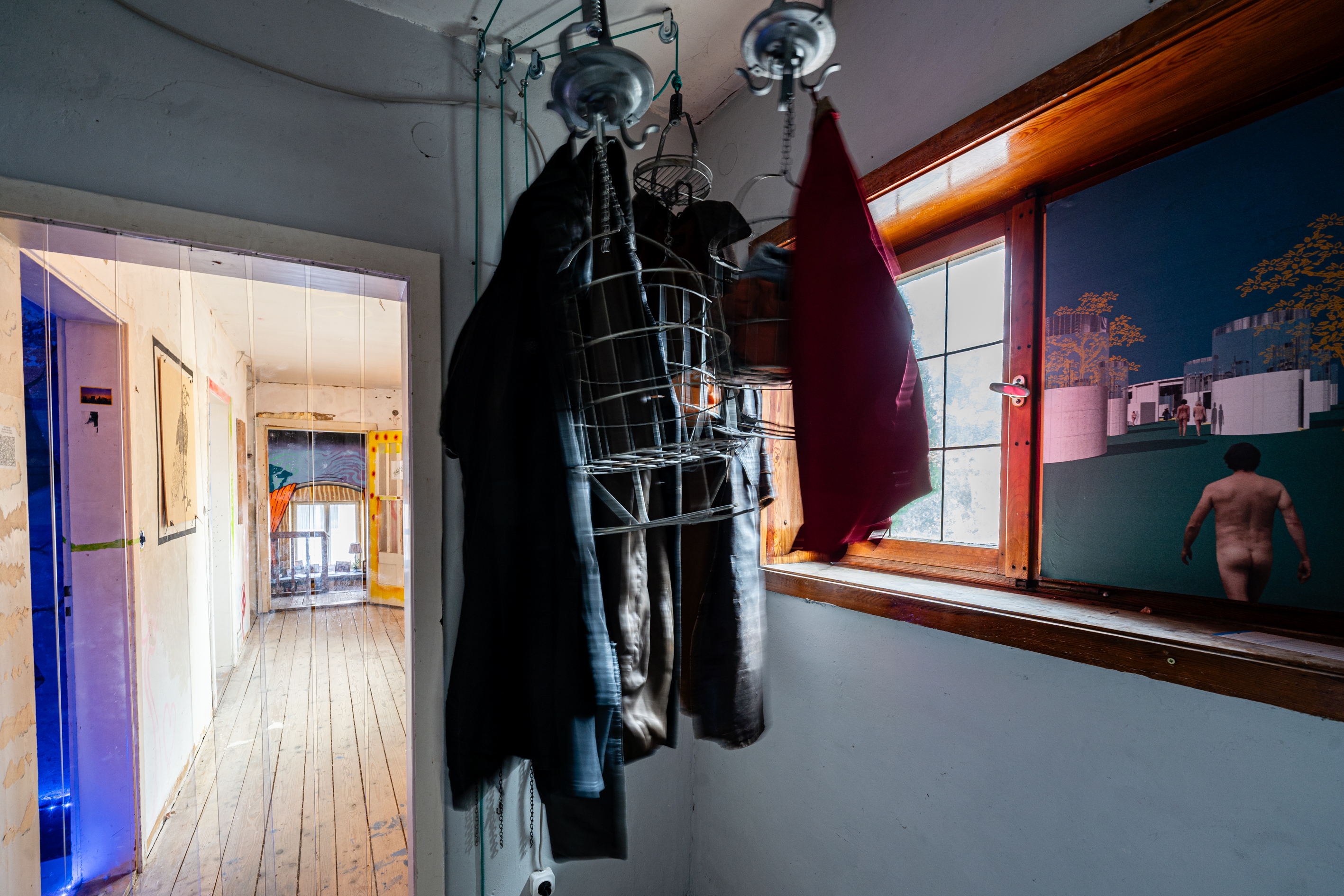 Bild 2 zu Die Welt als Fabrik: Blick auf eine Installation, die rechts neben der T&uuml;re h&auml;ngt und an der Decke mit drei Seilz&uuml;gen befestigt ist. An silbernen Haken h&auml;ngt Kleidung sowie Drahtk&ouml;rbe. An der rechten Wand befindet sich ein Fenster.