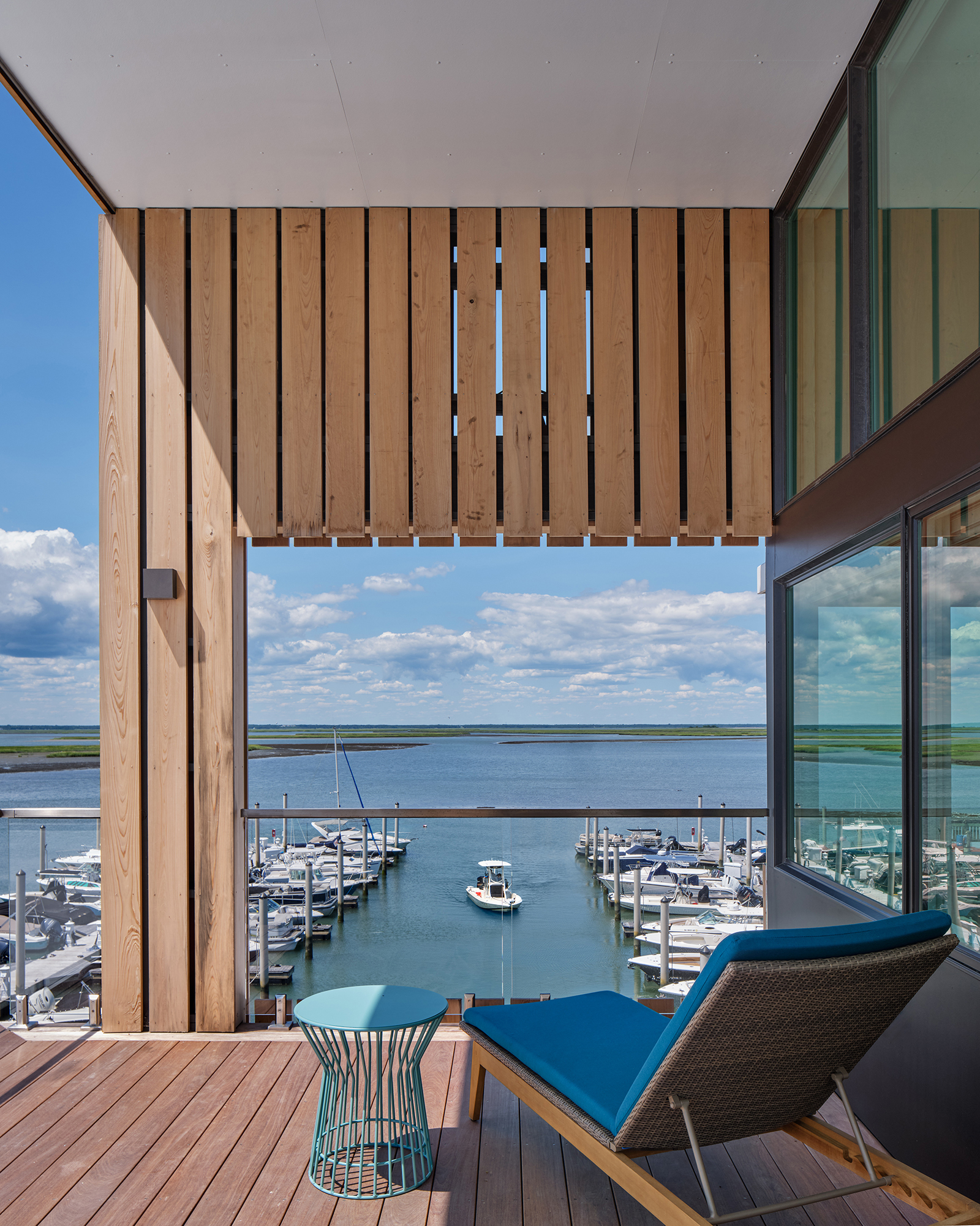 Interior and Exterior view from an upper-level deck, framed by vertical wood slats, showing a blue lounge chair and table, and a clear view down the marina channel to the open bay.