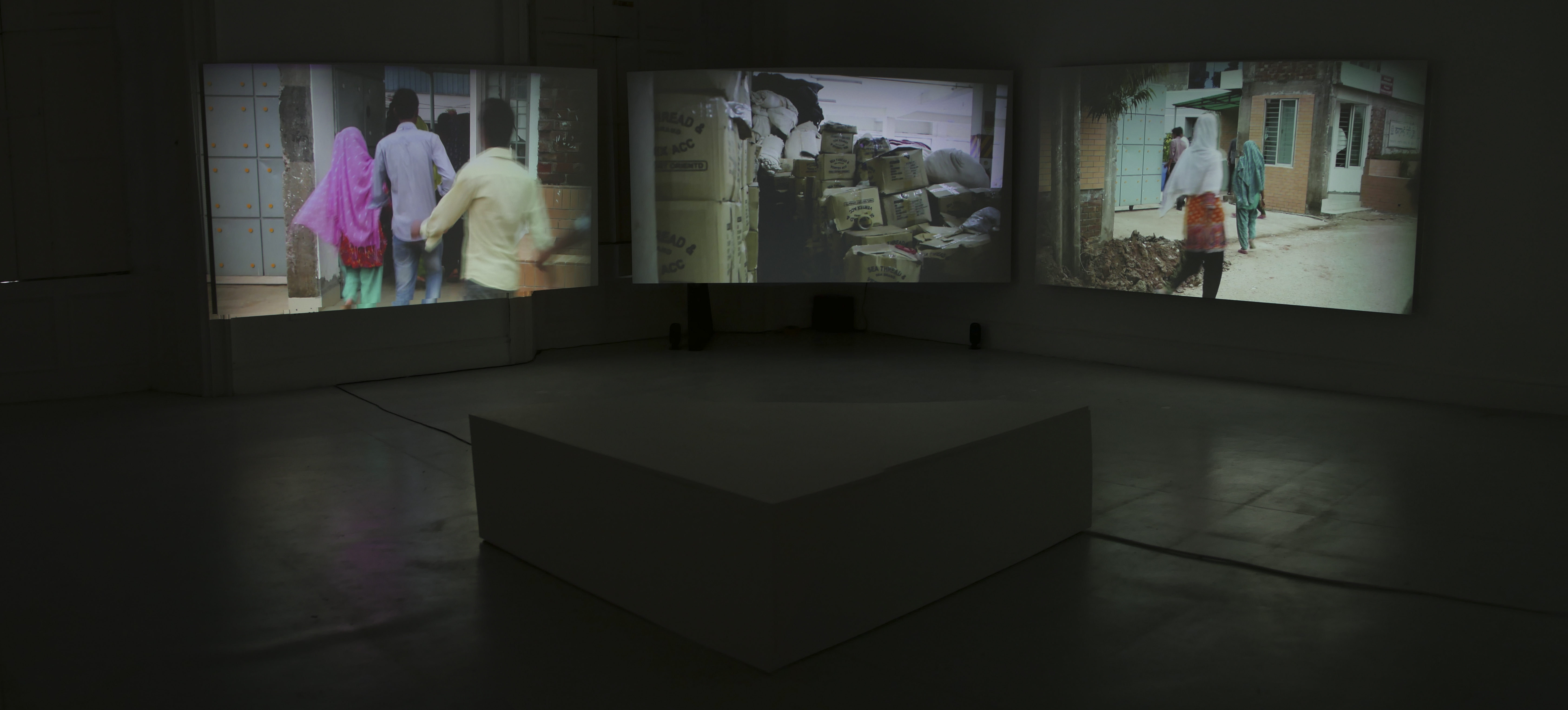 The image shows a white gallery space in low light. There are three screens mounted on the wall, each with a different image projected on it. From left to right the images show; workers entering a garment factory in Bangladesh; cardboard boxes full of completed garments piled high; workers leaving the garment factory via the front gate. In front on the three screens is a low bench for viewers to sit on.