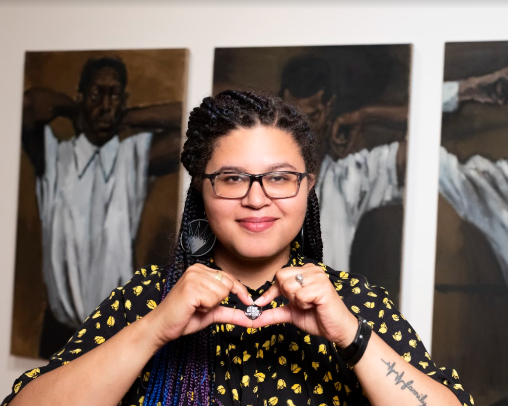 Esther Callahan, wearing a black button up short sleeve shirt shirt with a dotted yellow graphic flower pattern, glasses, and large hoop earrings, is looking directly at the camera in front of several larger than life portrait paintings, and is holding a small button with both hands in front of her which says “I am Black History.”