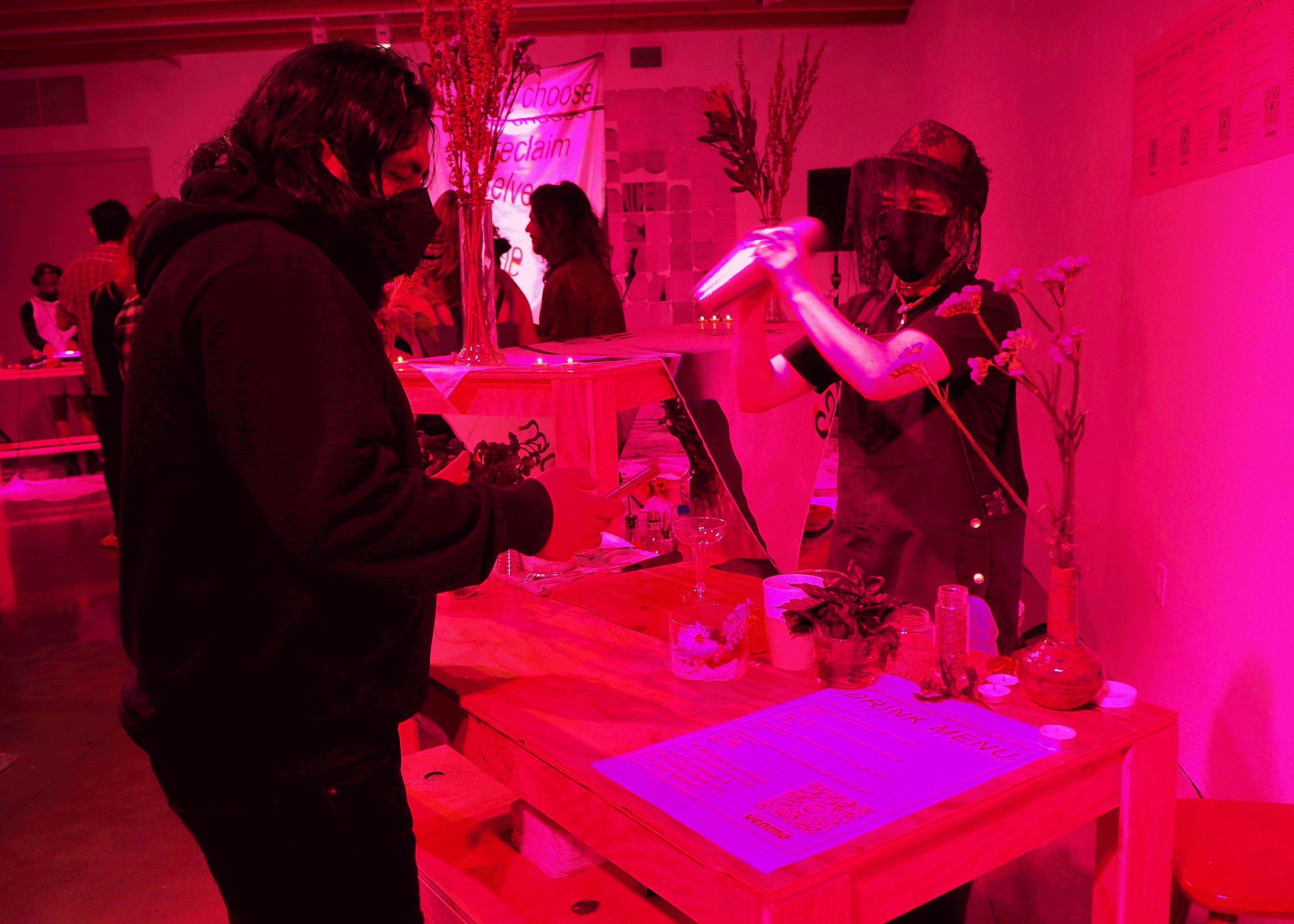 Under bright pink light, a masked and lace-veiled bartender shakes a metal cocktail shaker over their shoulder as a masked person dressed in black reviews the drink menu.